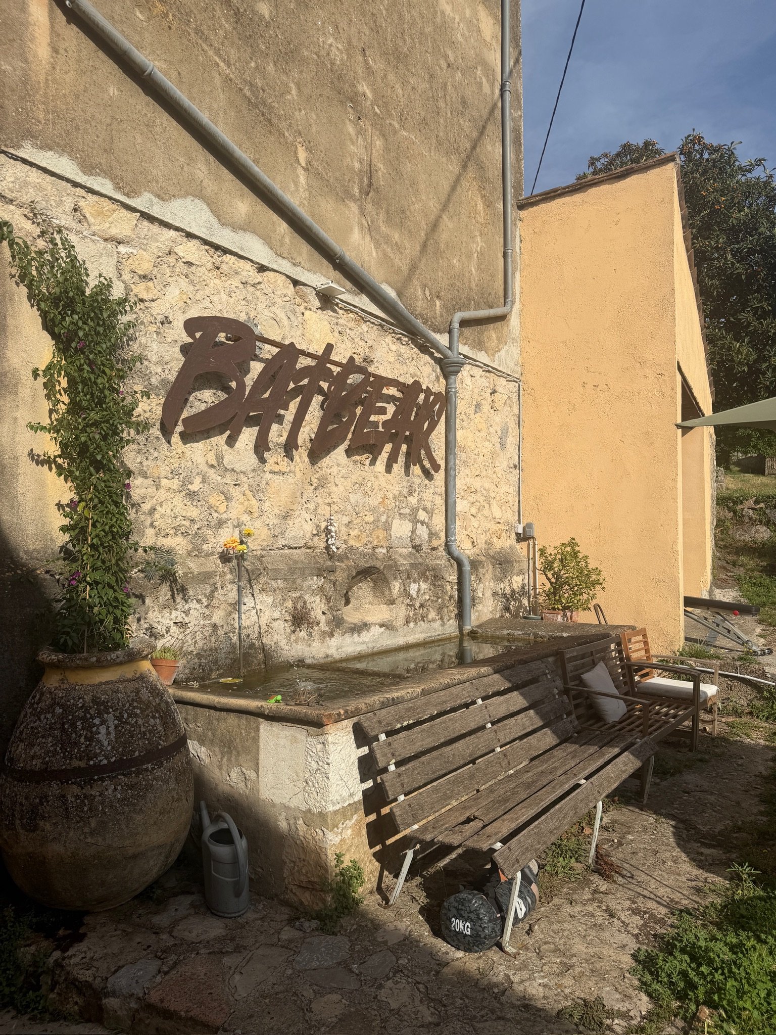 Une scène extérieure avec un mur en pierre et un mur en plâtre jaune, un banc en bois, un ballon de poids de 20 kg, un grand pot en terre cuite avec une plante grimpante, et une fontaine ou évier en pierre avec quelques herbes et fleurs.
