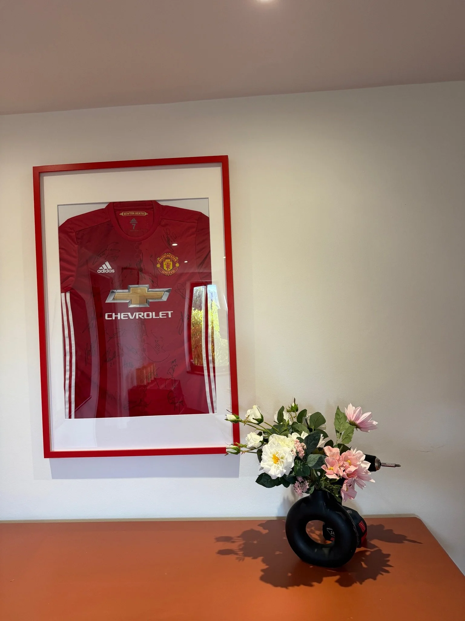 Cadre avec maillot de football Manchester United signé, suspendu au mur, à côté d'un bouquet de fleurs dans un vase noir sur une table en bois