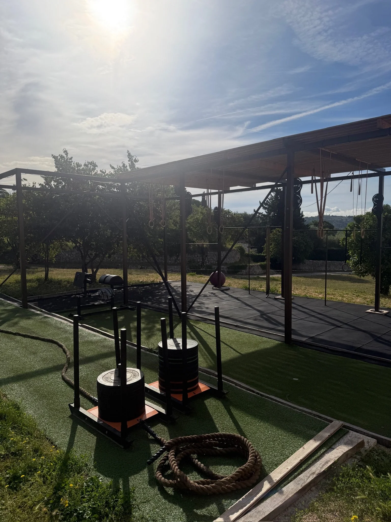Espace d'entraînement en plein air avec structure en métal, poids, cerceaux et corde, sous un ciel partiellement nuageux, entouré d'arbres et de végétation.