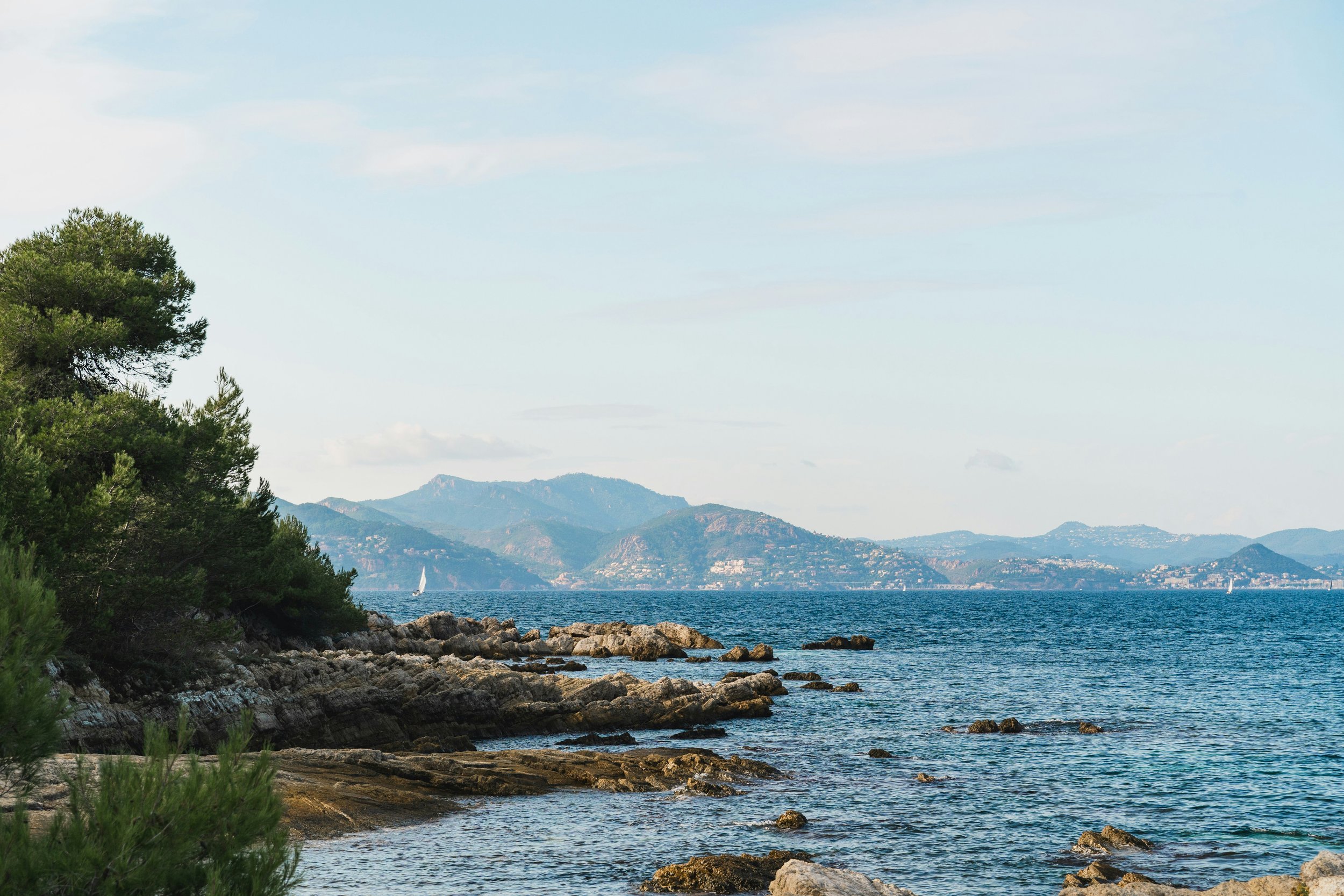 Côte rocheuse bordée d'arbres avec une mer calme et des montagnes en arrière-plan, sous un ciel en partie nuageux.
