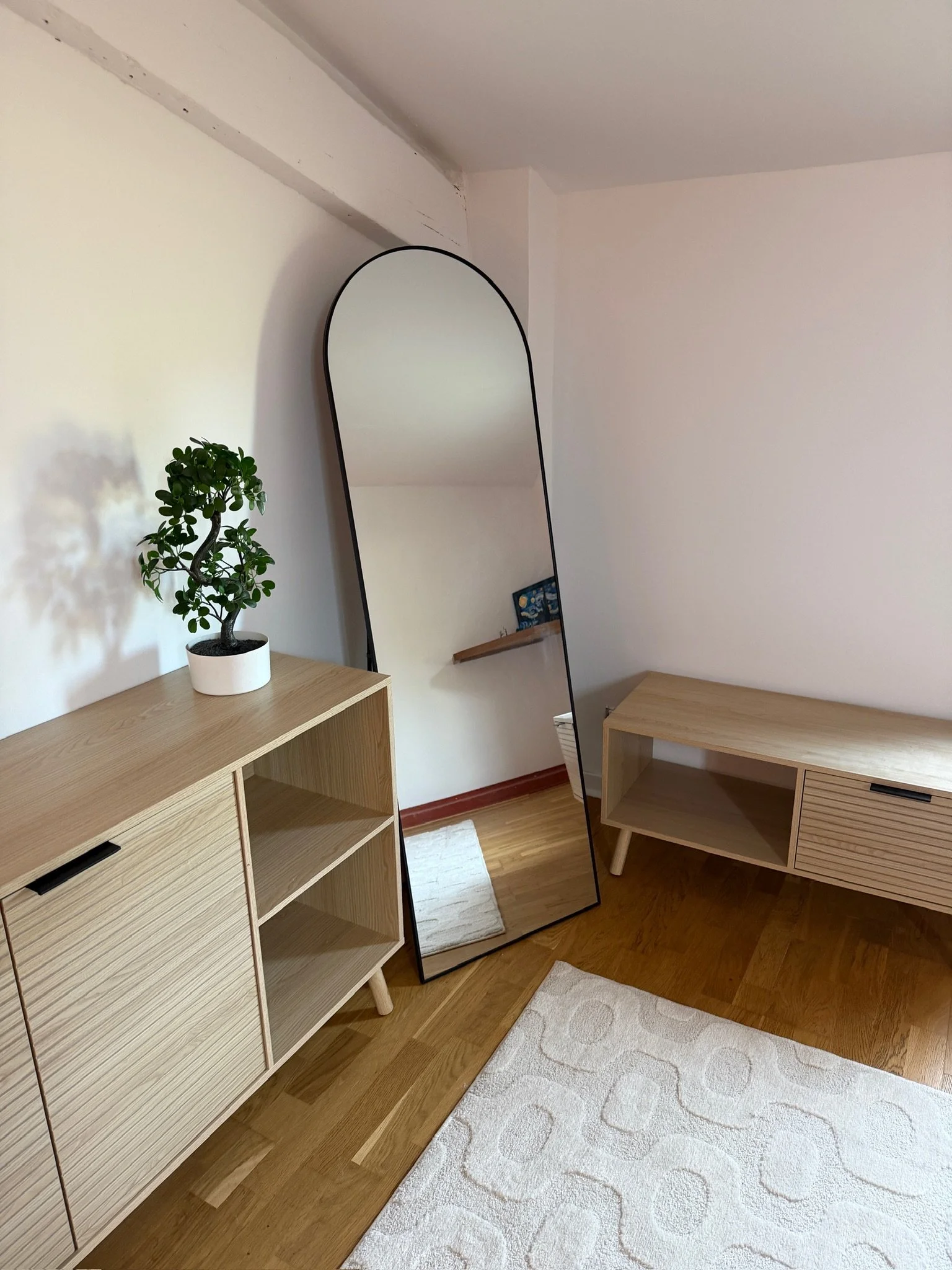 Interior de salón con un espejo de pie, un mueble bajo de madera clarísima con puertas de listones y un armario alto de madera con puertas de listones, sobre una alfombra blanca con patrón circular, en una esquina, una planta en maceta blanca.