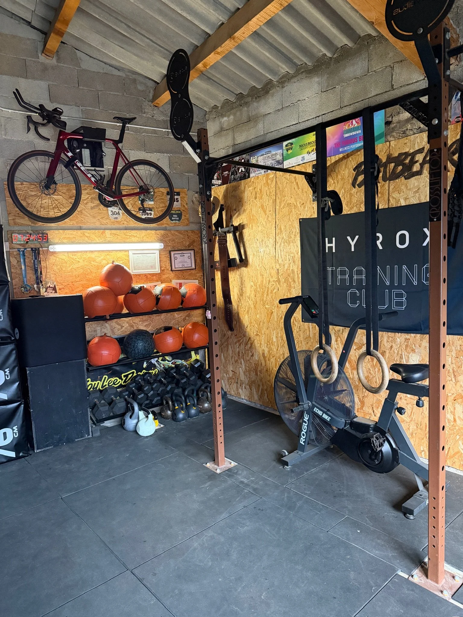 Salle de sport avec tapis de course, haltères, jeux de poids, ballons de médecine, et un mur avec des affiches et un chronomètre numérique.