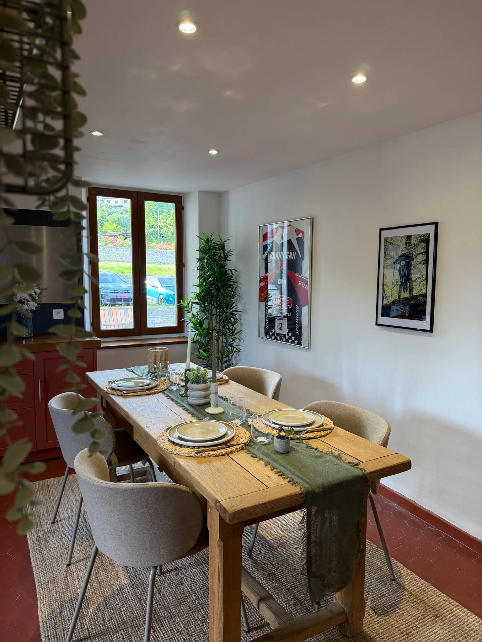 Une salle à manger avec un grand table en bois, six chaises beiges, assiettes, verres et plantes décoratives. Un mur avec des affiches, une fenêtre laissant entrer la lumière.