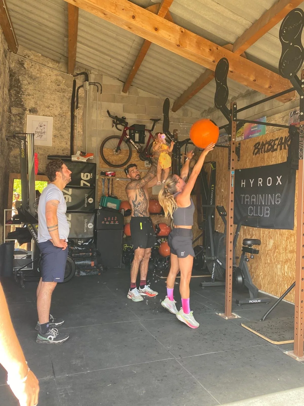 Groupe de personnes dans une salle de sport effectuant un entraînement. Deux femmes sautent pour attraper un ballon orange, un homme avec un tatouage les regarde. Un autre homme et une autre personne aussi regardent l'activité. On peut voir des équip
