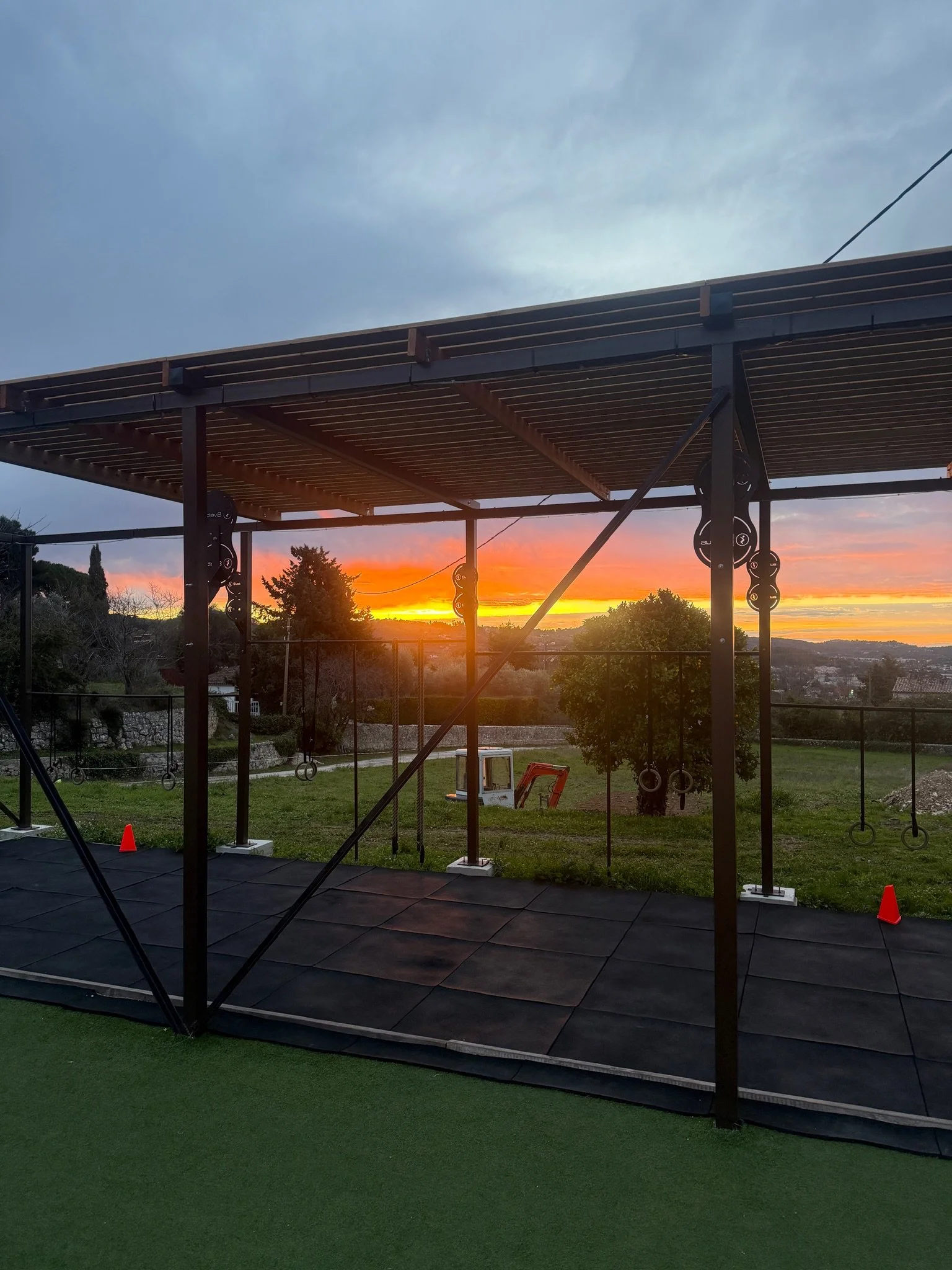 Une structure de salle d'entraînement en plein air avec un toit en bois, des anneaux de gymnastique et une peinture en gazon vert sur le sol, avec un coucher de soleil en arrière-plan