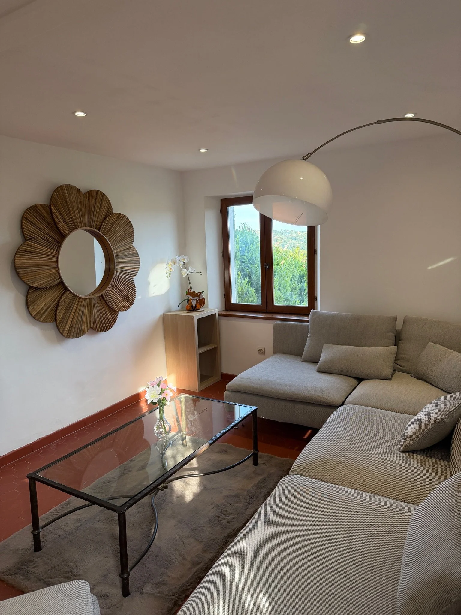 Salon avec canapé gris, table en verre, mur blanc, miroir en forme de fleur en bois, fenêtre avec vue sur la verdure, lampe suspendue blanche, petite étagère avec décoration de cerf en bois et fleurs