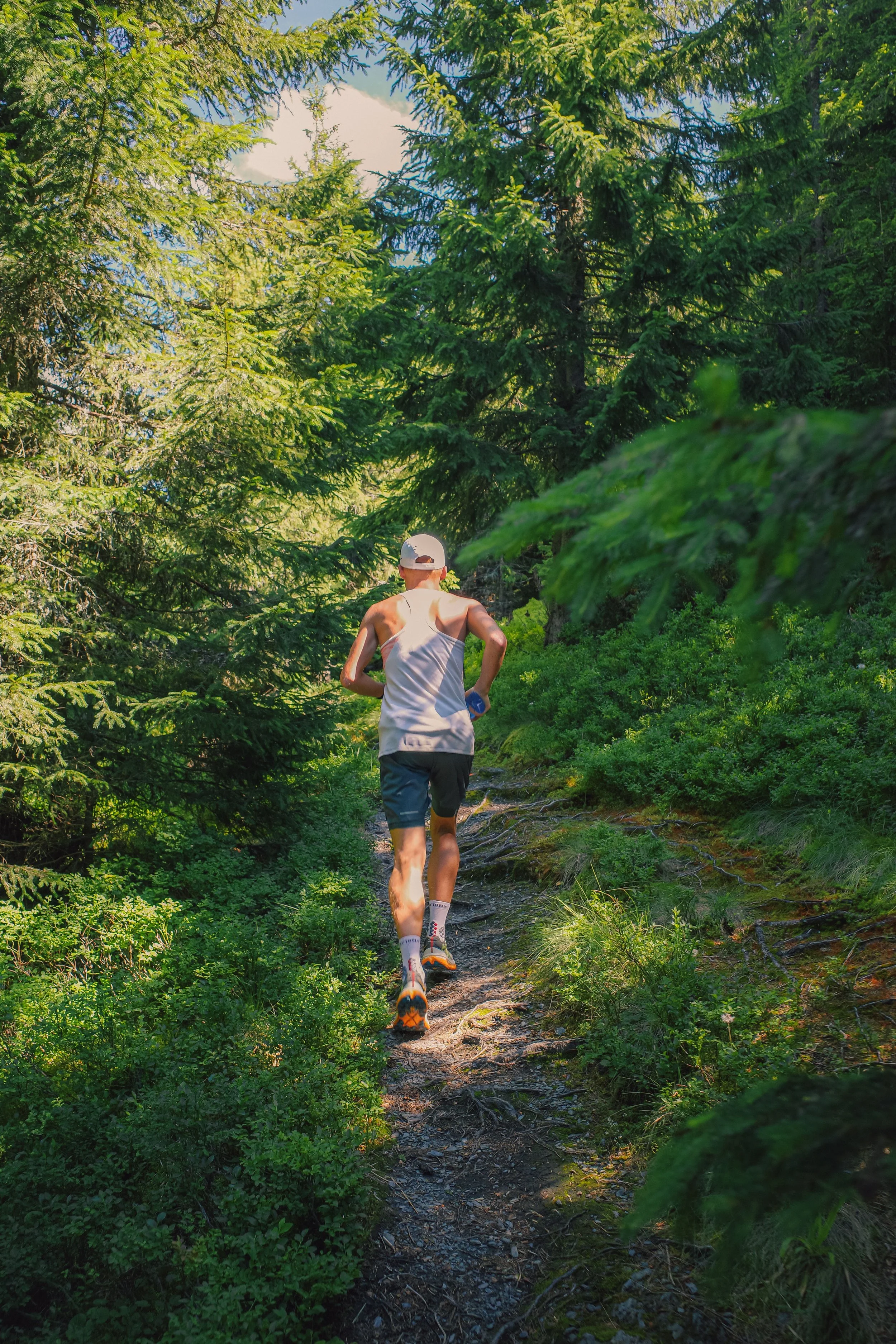 Un homme court sur un sentier dans une forêt dense en été, portant un débardeur blanc, un casque blanc, des shorts gris et des chaussures de course orange et noir.