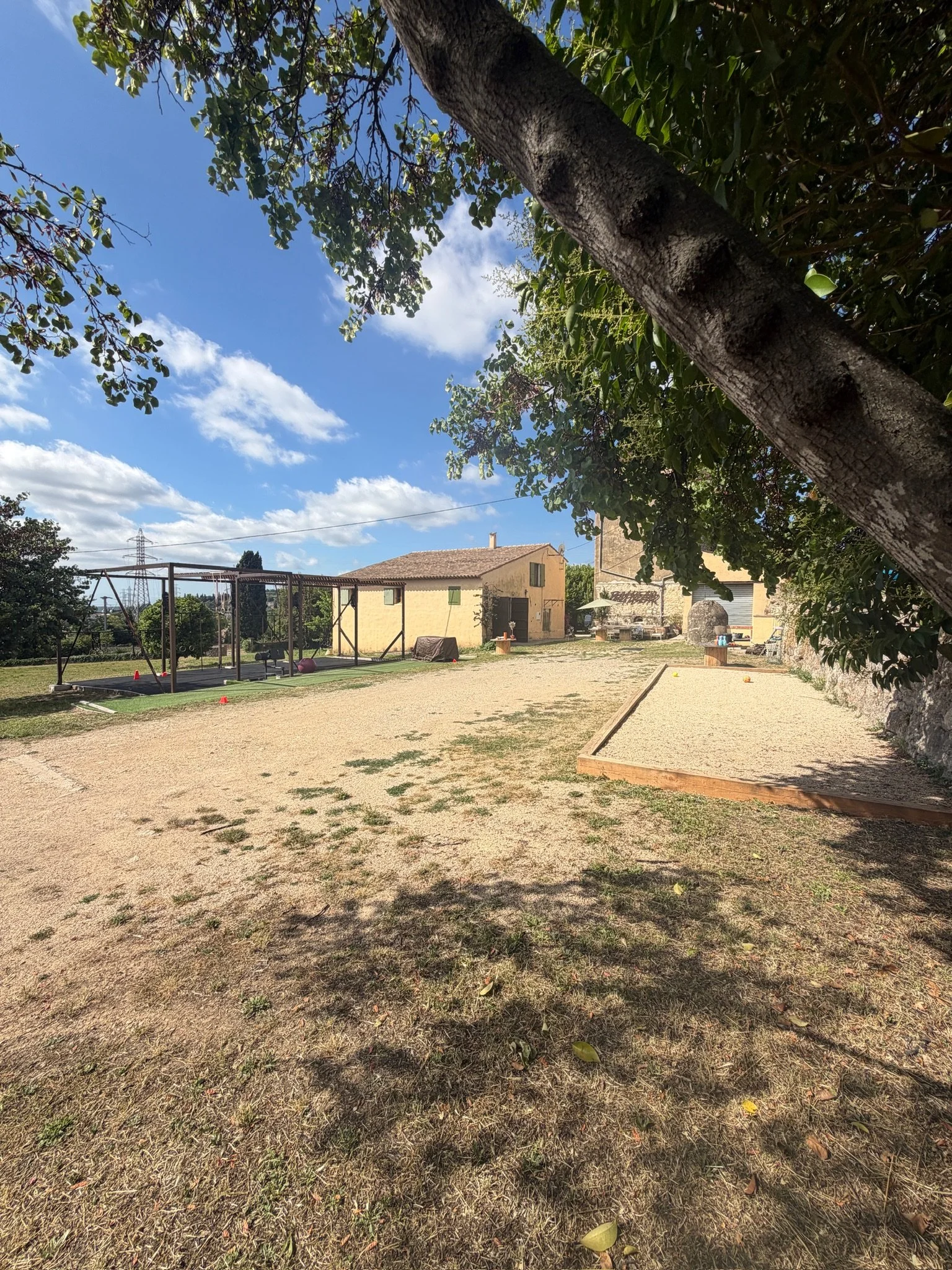 Un espace de jeu pour enfants avec une balançoire, un trampoline, une maison en pierre, un grand arbre, un sol recouvert de sable et un ciel bleu avec quelques nuages, dans un jardin.Enfants jouent dans le jardin par une journée ensoleillée.
