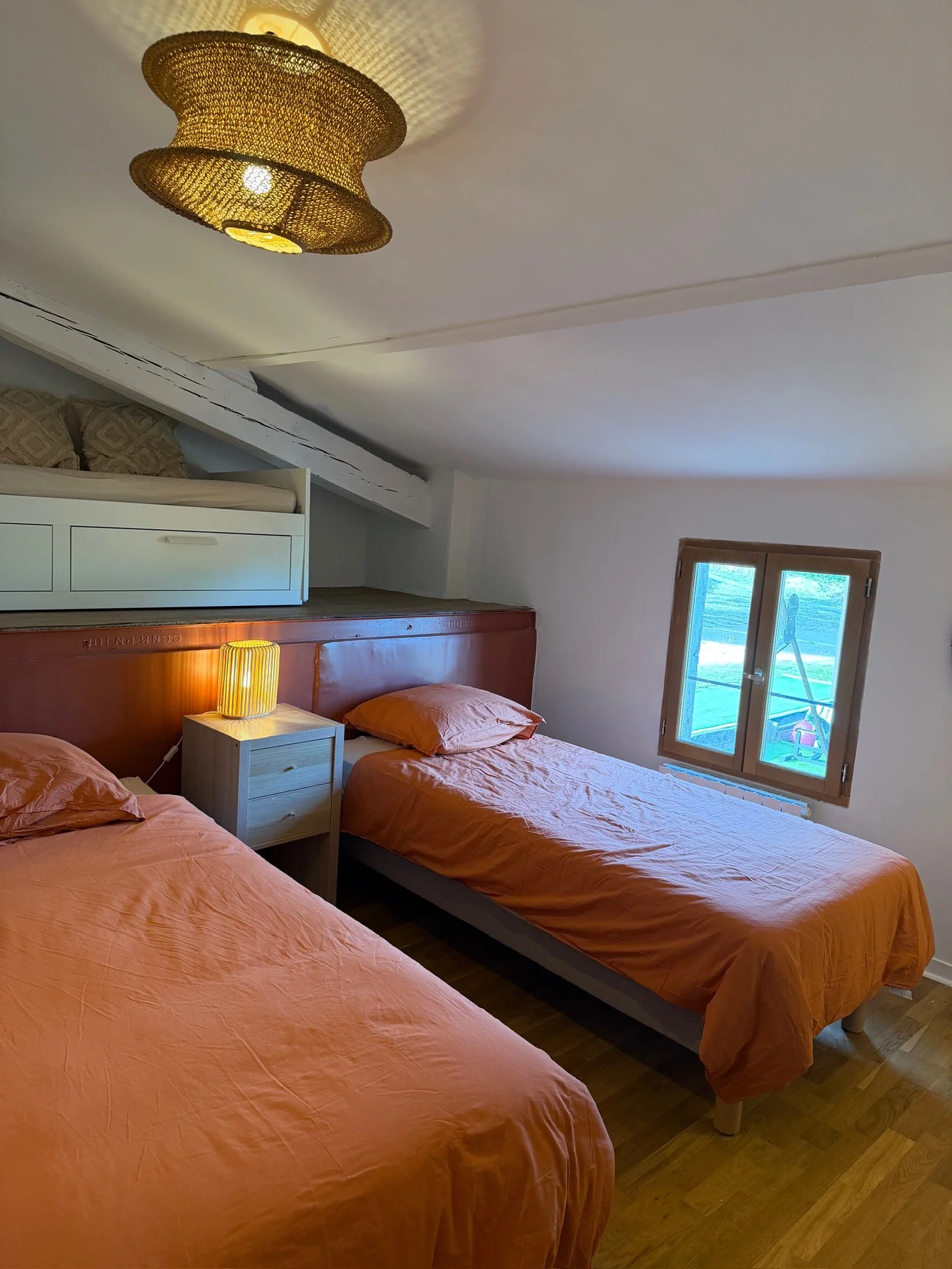 Chambre avec trois lits simples en orange, chevet avec lampe, fenêtre en bois, plafond incliné, déco minimaliste, ambiance chaleureuse.