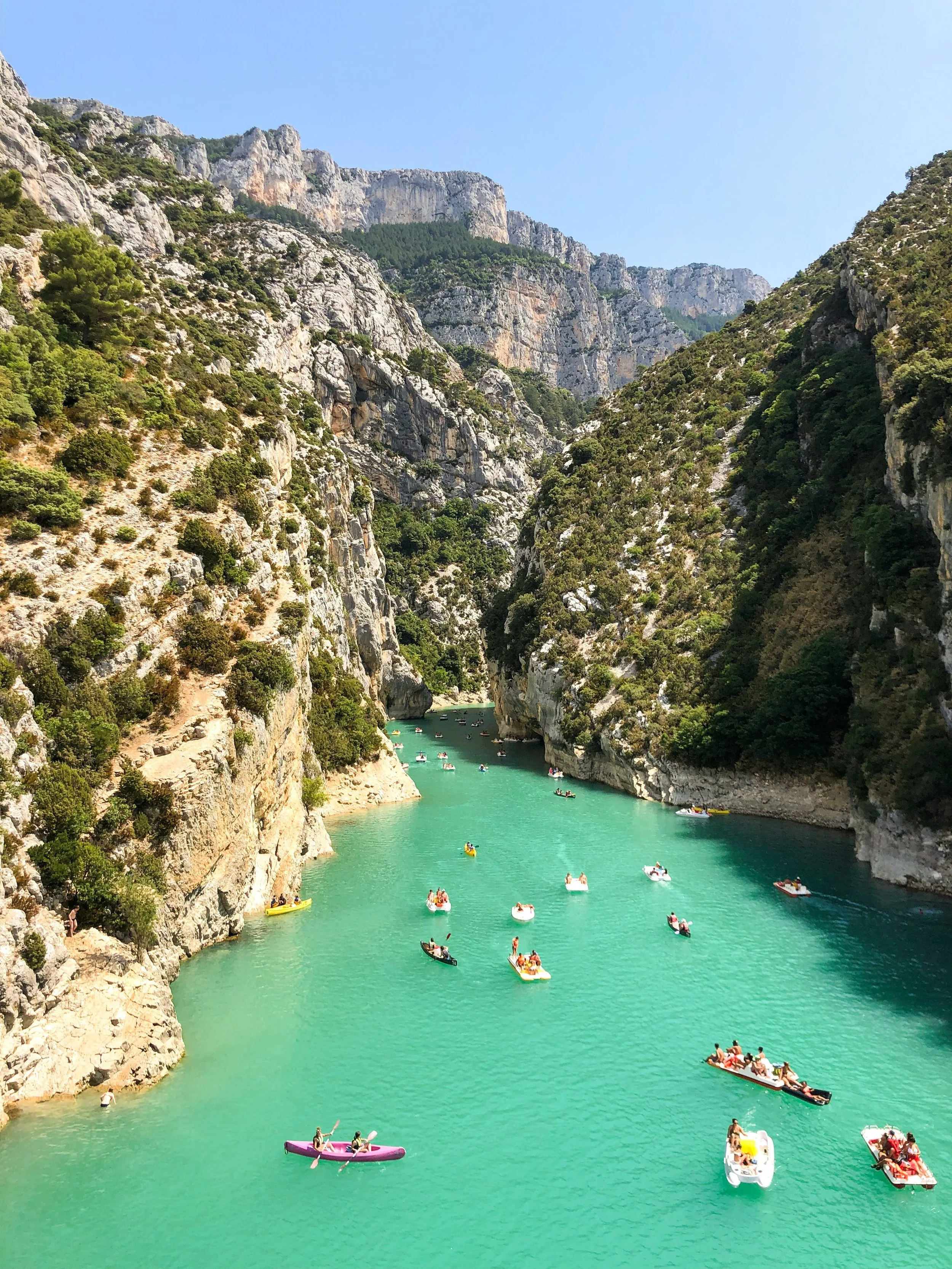 Une rivière turquoise entourée de falaises rocheuses et de verdure, avec des bateaux et des kayaks flottant sur l'eau, sous un ciel bleu.