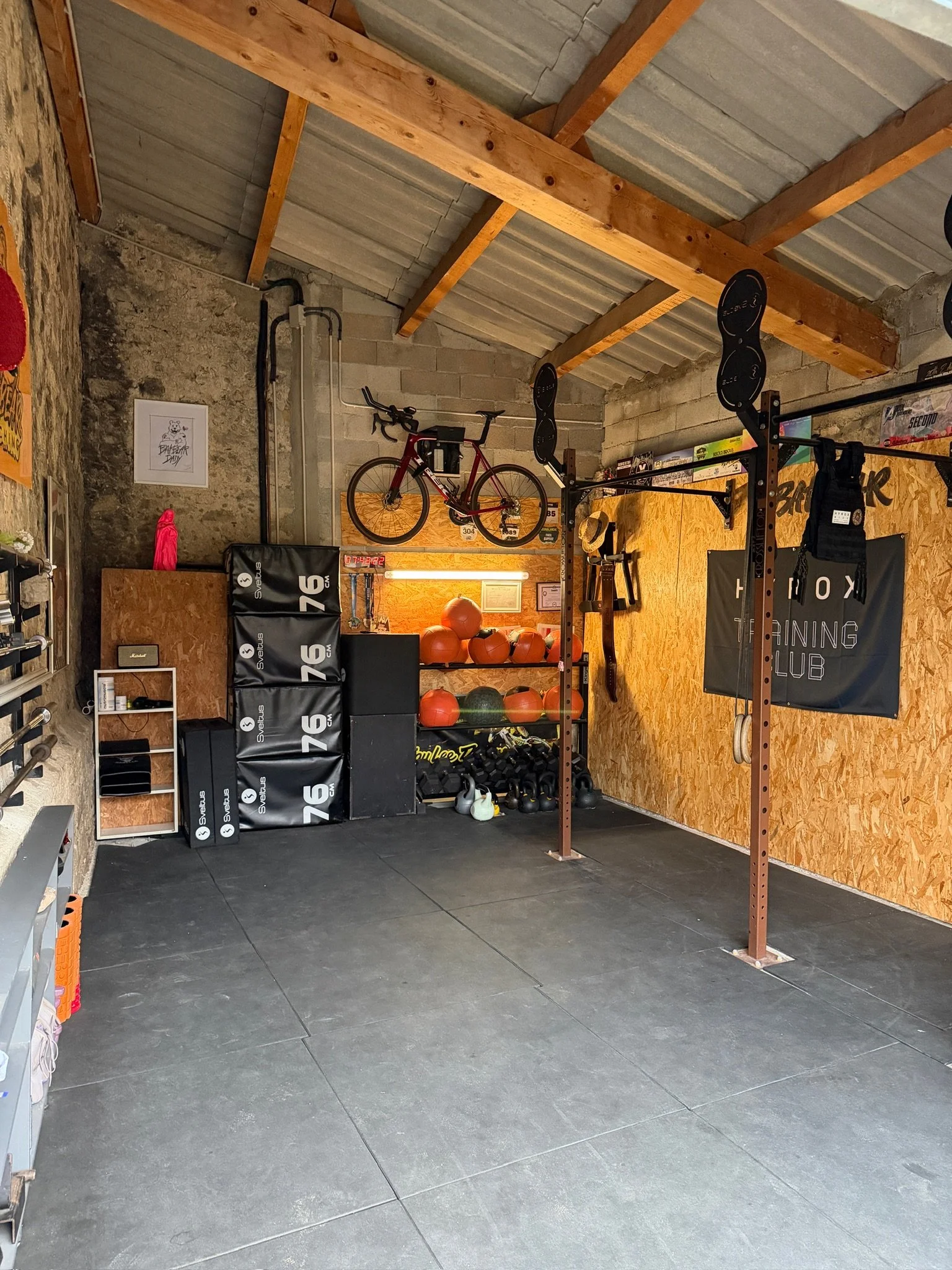 Salle de sport ou salle d'entraînement avec équipements de fitness, un vélo fixé en haut, sacs de pliométrie, ballons de médecine, haltères et accrochements pour barres de musculation sur un mur en bois, et un mur en pierre visible à gauche.