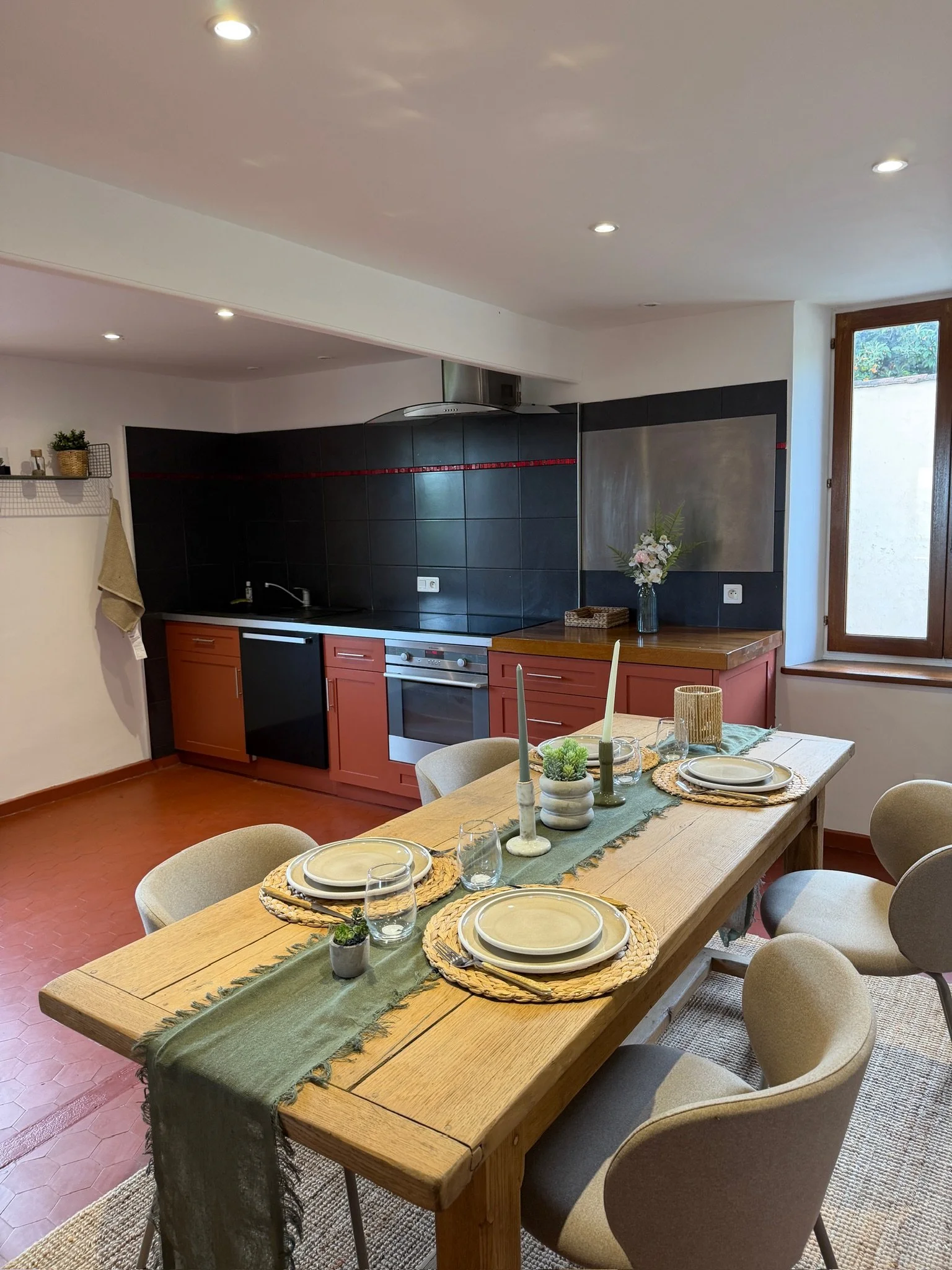 Salle à manger avec une grande table en bois, décorée de vaisselle, de bougies, de plantes en pot et d'une nappe verte. En arrière-plan, une cuisine avec des armoires rouges, un plan de travail noir et une fenêtre laissant entrer la lumière.