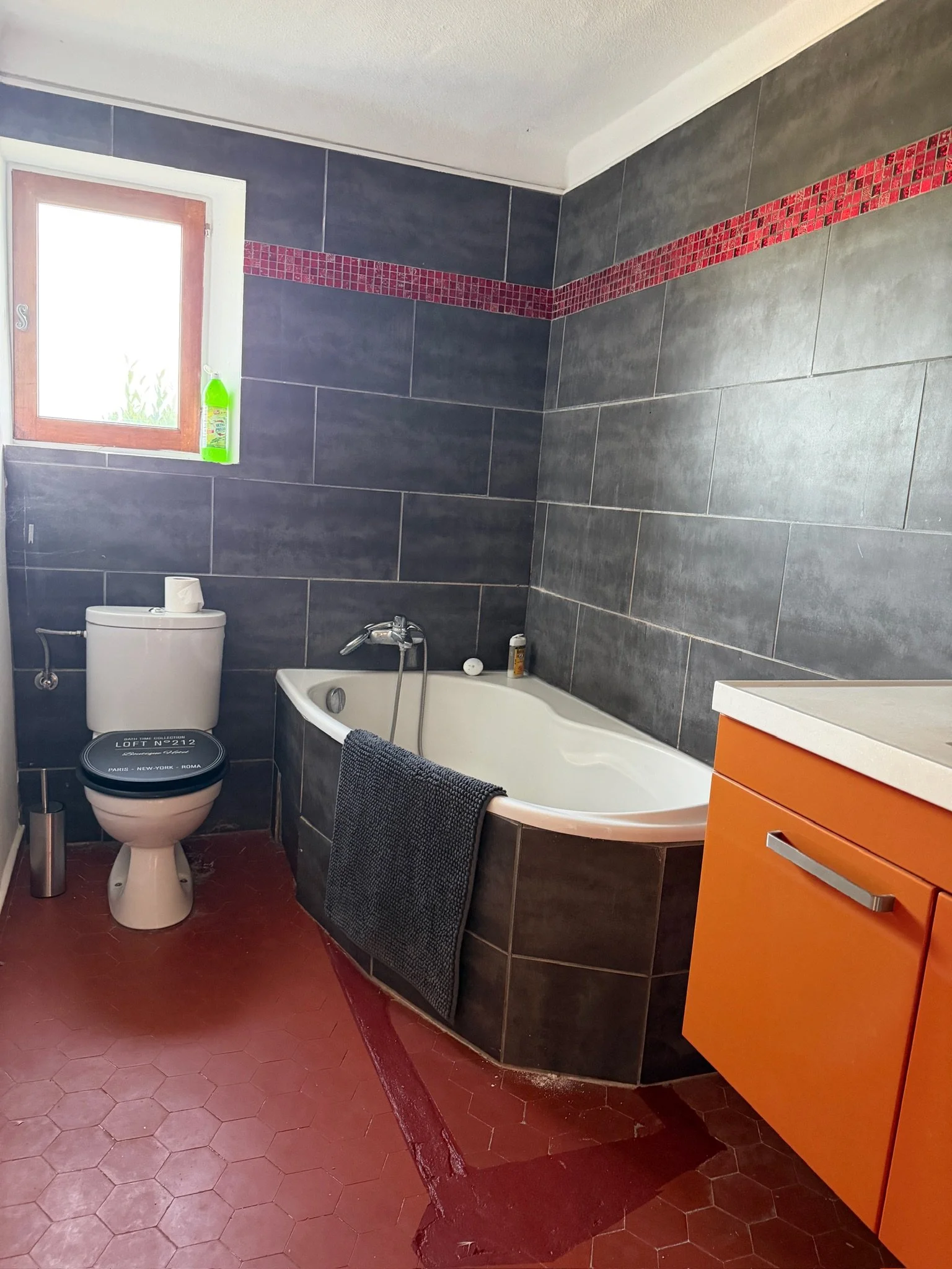 Une salle de bain avec murs en carrelage gris, une baignoire, un toilette avec un rouleau de papier toilette, une fenêtre avec une bouteille de nettoyant, et un meuble orange avec tiroirs.