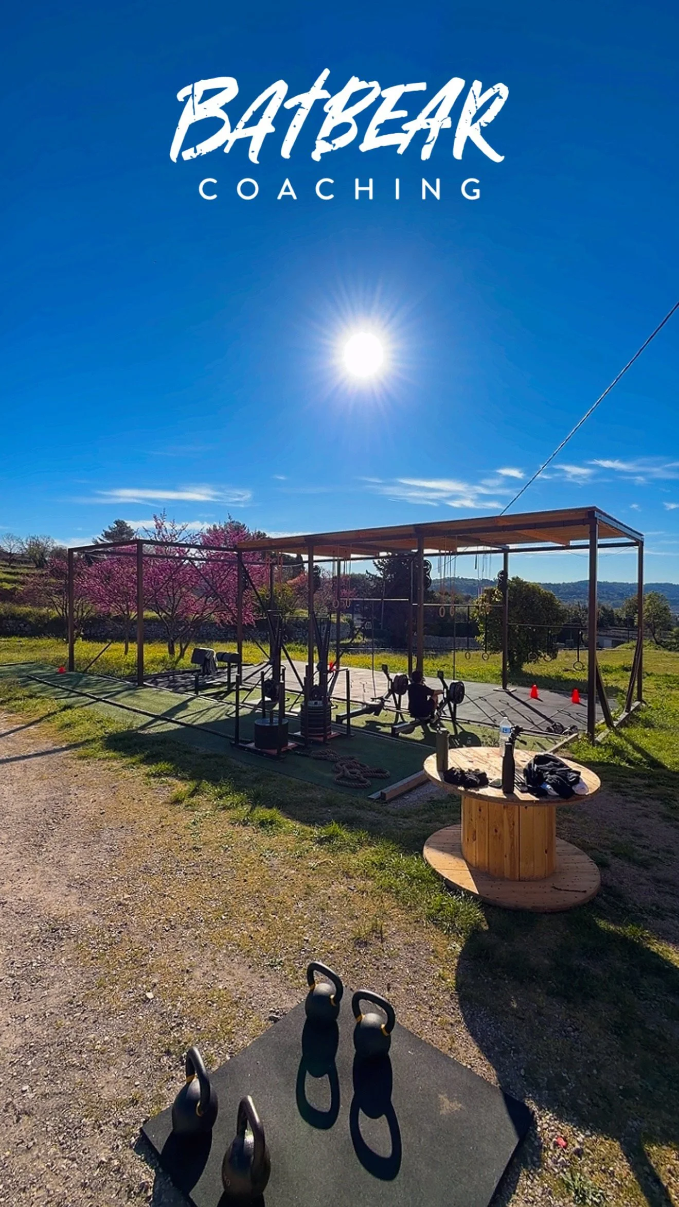 Une salle d'entraînement extérieure avec des équipements de musculation, un tapis, un câble de poulie, un tableau en bois, un terrain herbeux, des arbres en fleur et un ciel ensoleillé.