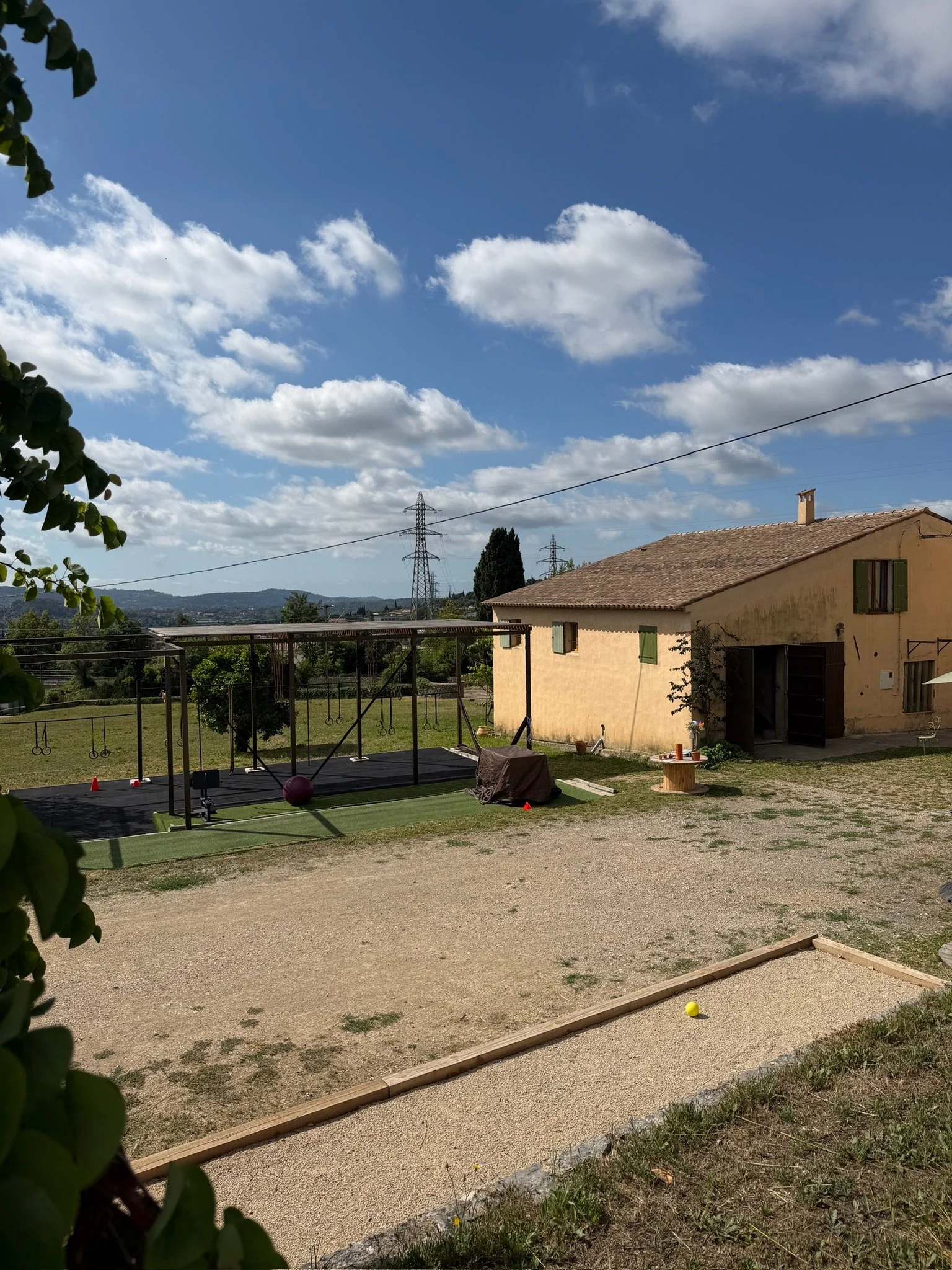 Un jardin avec une maison beige, un terrain de sport, des arbres, des poteaux électriques, un ciel bleu avec des nuages blancs.