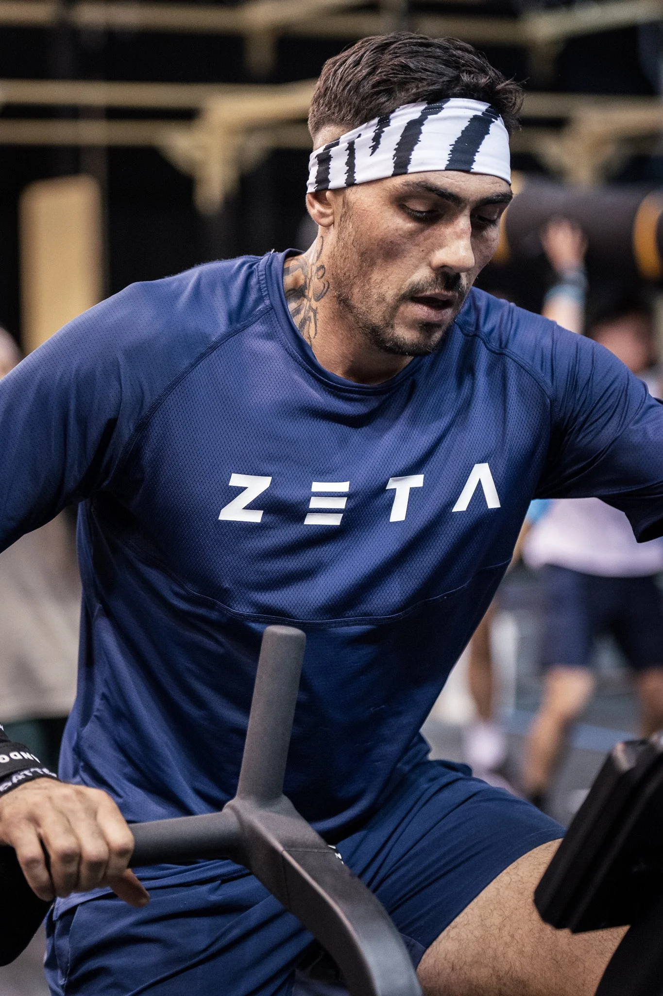 Le coach sportif faisant de l'exercice sur un vélo elliptique, portant un t-shirt bleu avec l'inscription 'Z.E.T.A' et un bandeau noir et blanc, concentré pendant l'entraînement.