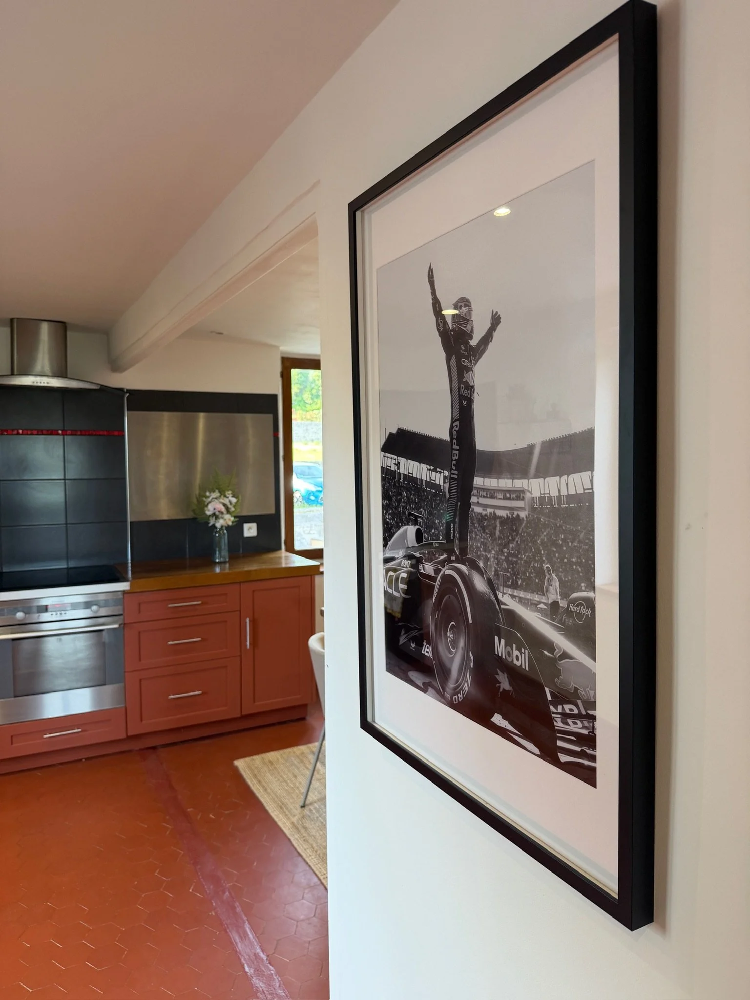 Photo d'une pièce intérieure avec un tableau en noir et blanc représentant une victoire de Formule 1, accroché à un mur blanc. À l'arrière-plan, une cuisine avec des meubles en bois rouge et un vase de fleurs.