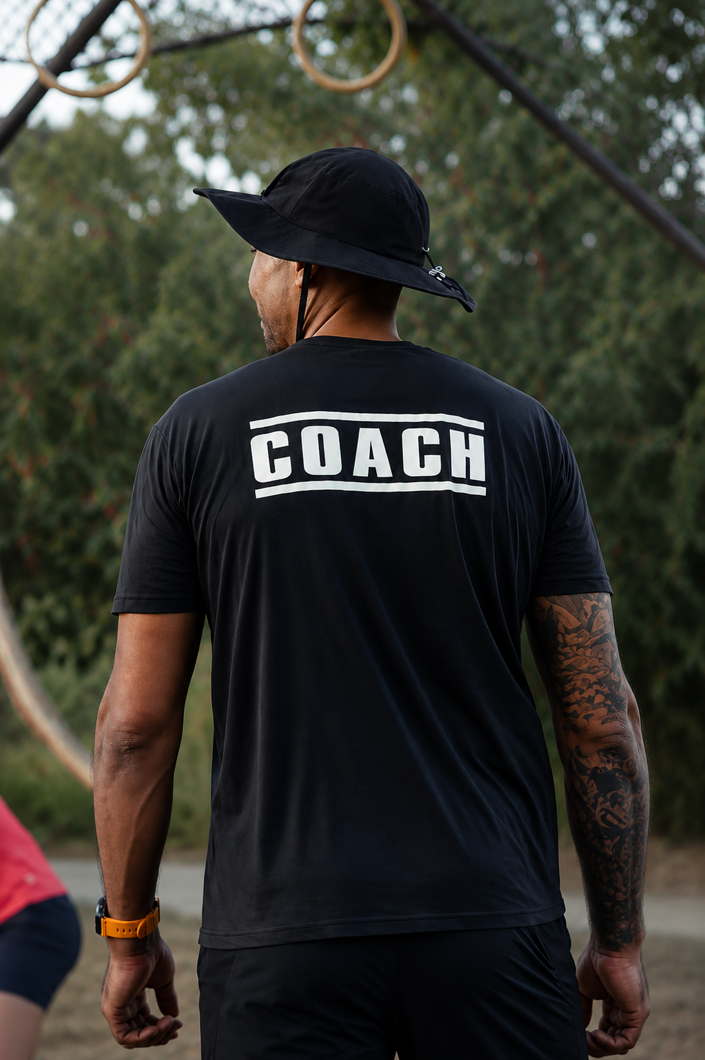 Homme vu de dos portant un t-shirt noir avec le mot "COACH" imprimé en blanc dans le dos, un chapeau noir, une montre orange et un bras tatoué, dans un parc avec des arbres et des anneaux de gymnastique en arrière-plan.