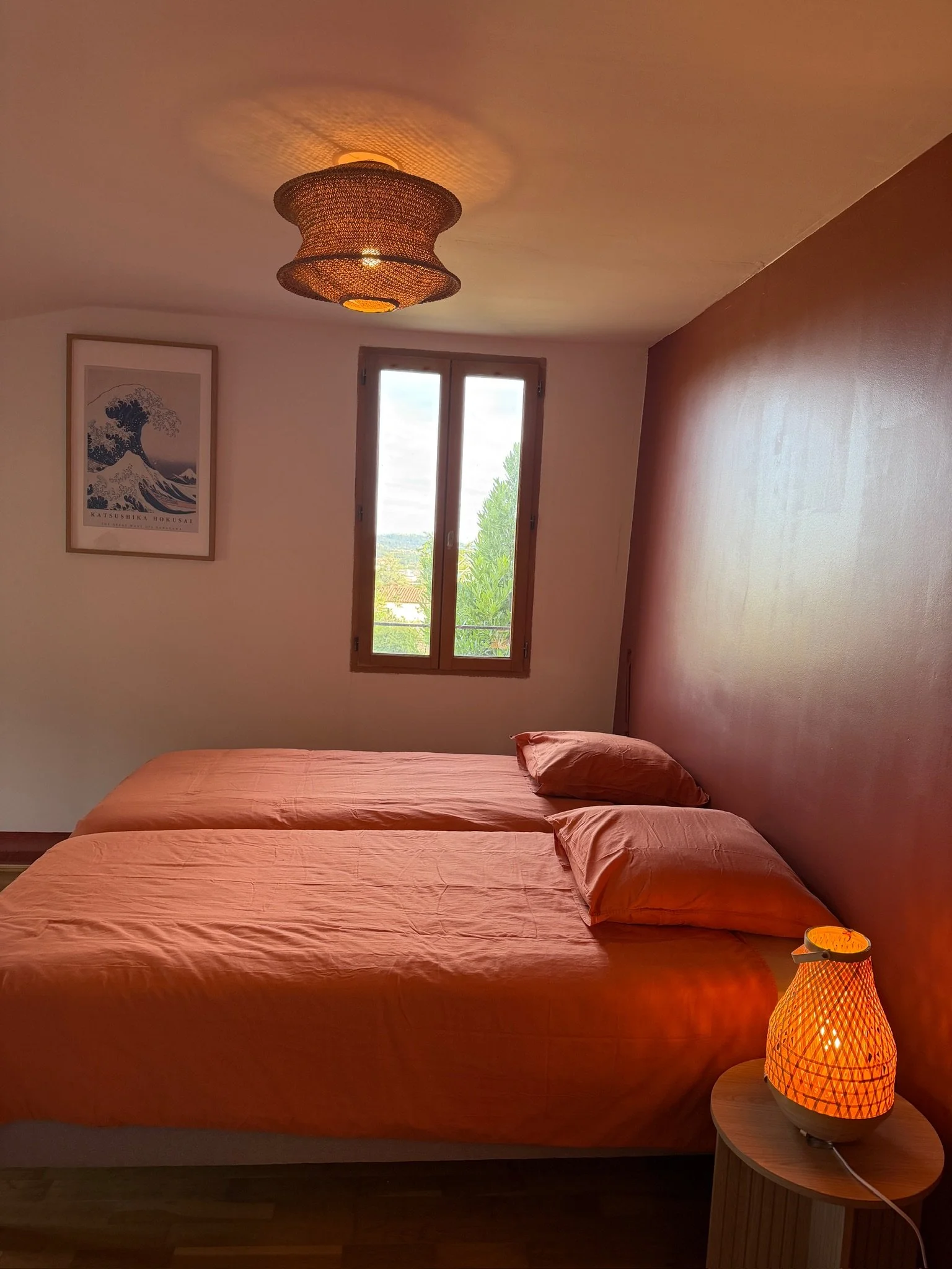 Une chambre avec deux lits orangés, une fenêtre, un tableau représentant une vague de Hokusai, une lampe de chevet en osier émettant une lumière chaude.