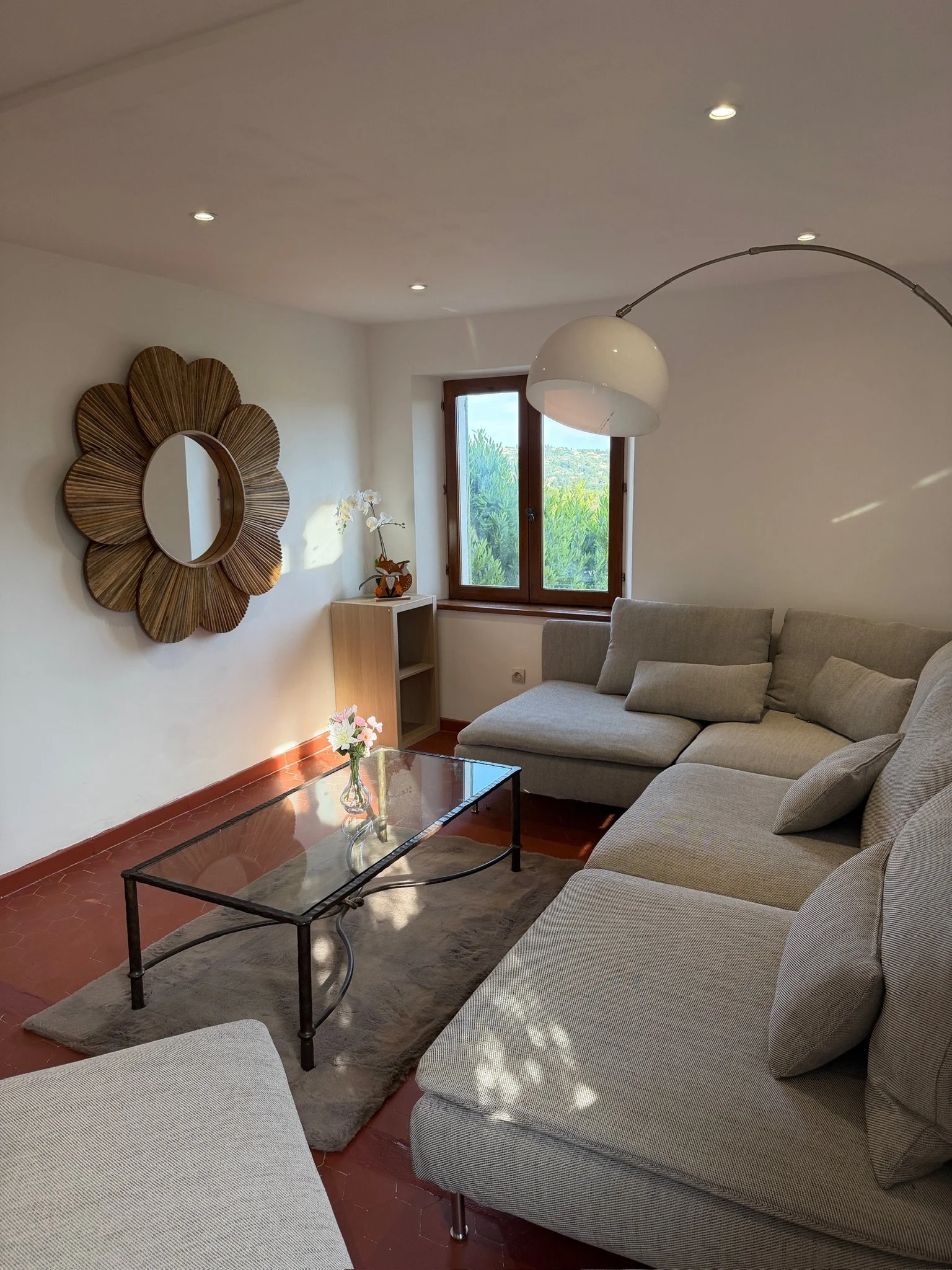 Salon avec un canapé gris, une table en verre, un miroir en forme de fleur en bois, une fenêtre, une lampe en arc, et une décoration florale sur une petite table.