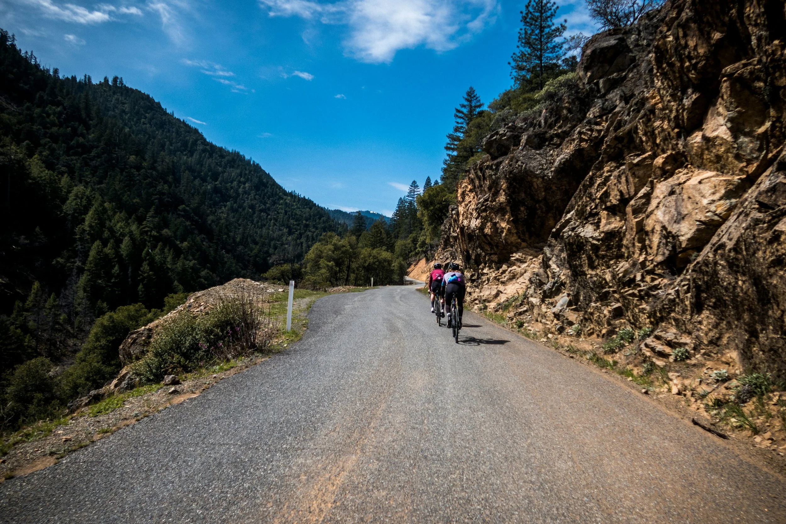 Trois cyclistes pédalant sur une route de montagne entourée de falaises rocheuses et de forêts verdoyantes, sous un ciel bleu avec quelques nuages.