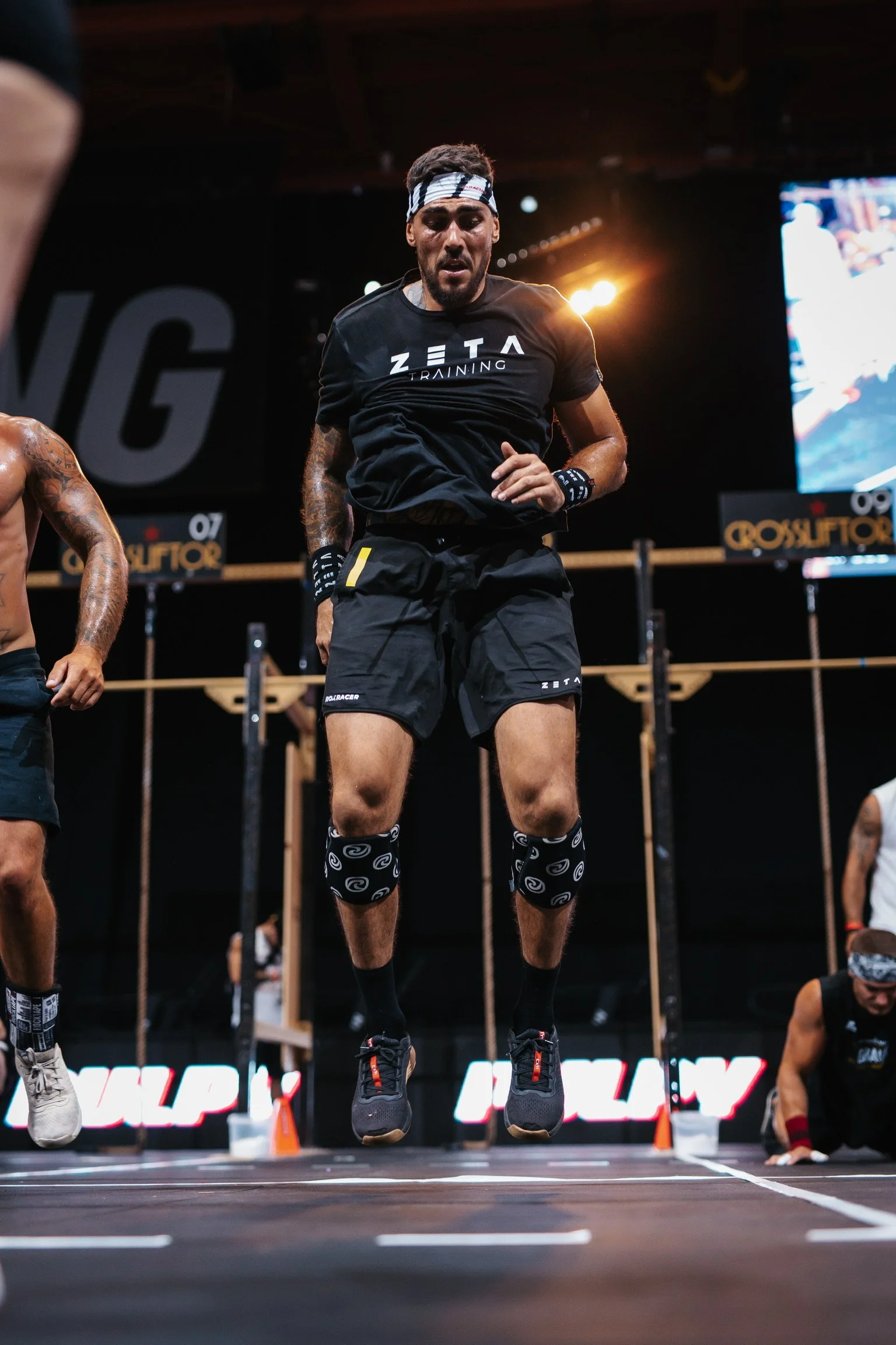 Un homme en tenue de sport effectuant un saut lors d'une compétition de CrossFIt en intérieur, portant un bandeau blanc et noir, une t-shirt noir avec le logo Zeta Training, shorts noirs, et des genouillères noires et blanches.