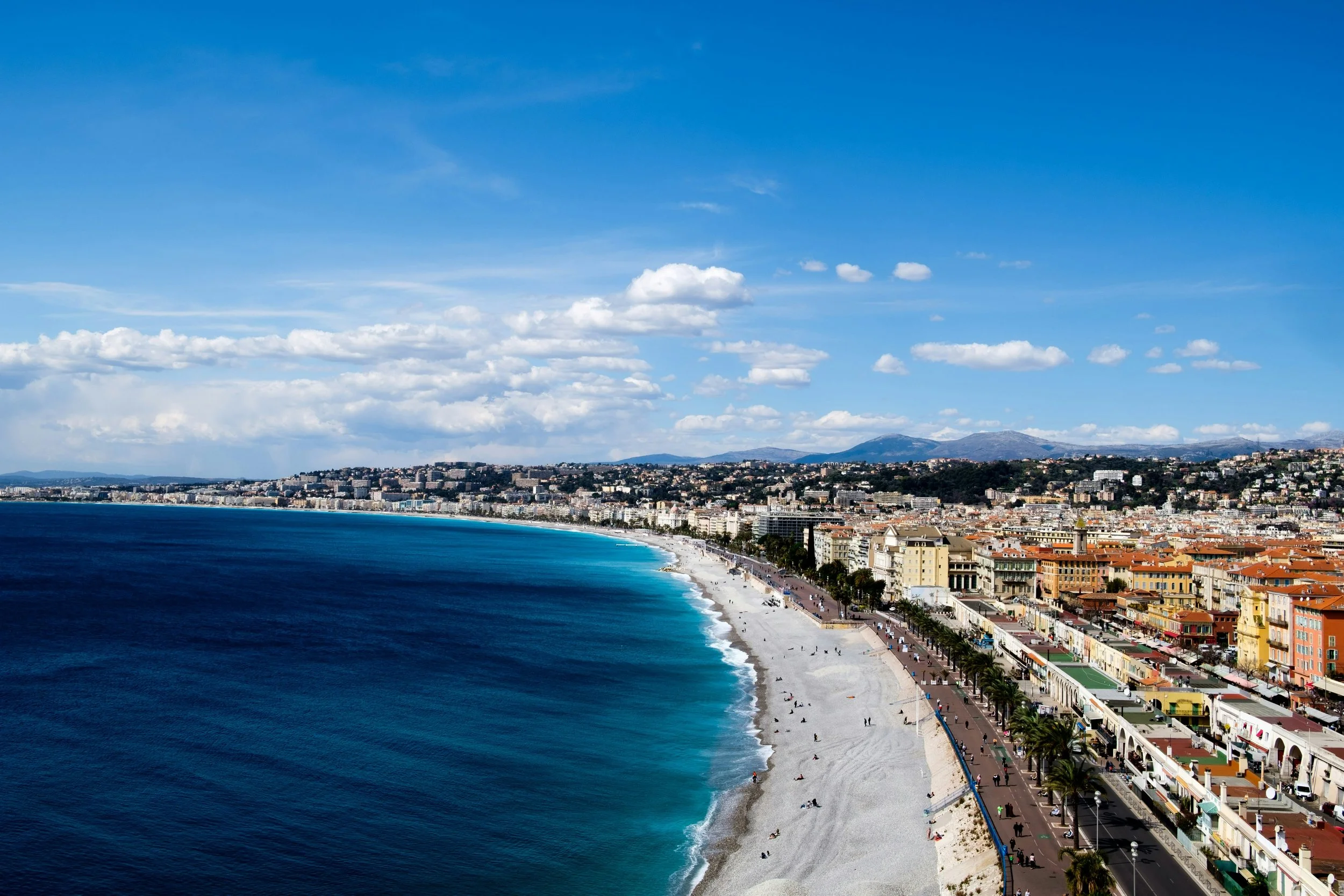 Une vue aérienne d'une plage urbaine avec des bâtiments colorés et une promenade, bordée par la mer avec des vagues calmes, sous un ciel bleu avec quelques nuages.
