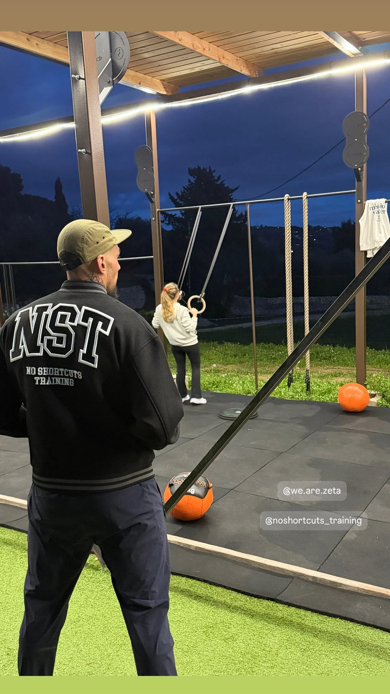 Un entraîneur observe deux personnes en entraînement de musculation ou de fitness en extérieur, avec des équipements comme des cordes, une balle de médecine, et un sac de sable, lors d'une séance en soirée sous un ciel nocturne.