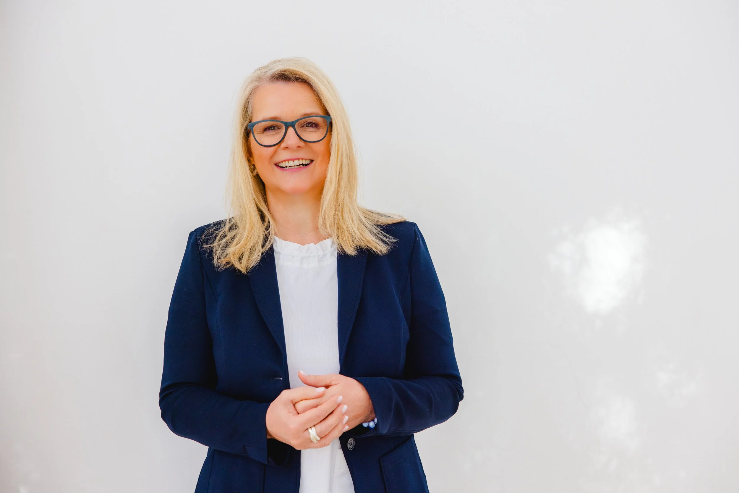 Lächelnde blonde Frau mit Brille in dunkler Blazerjacke vor weißer Wand.