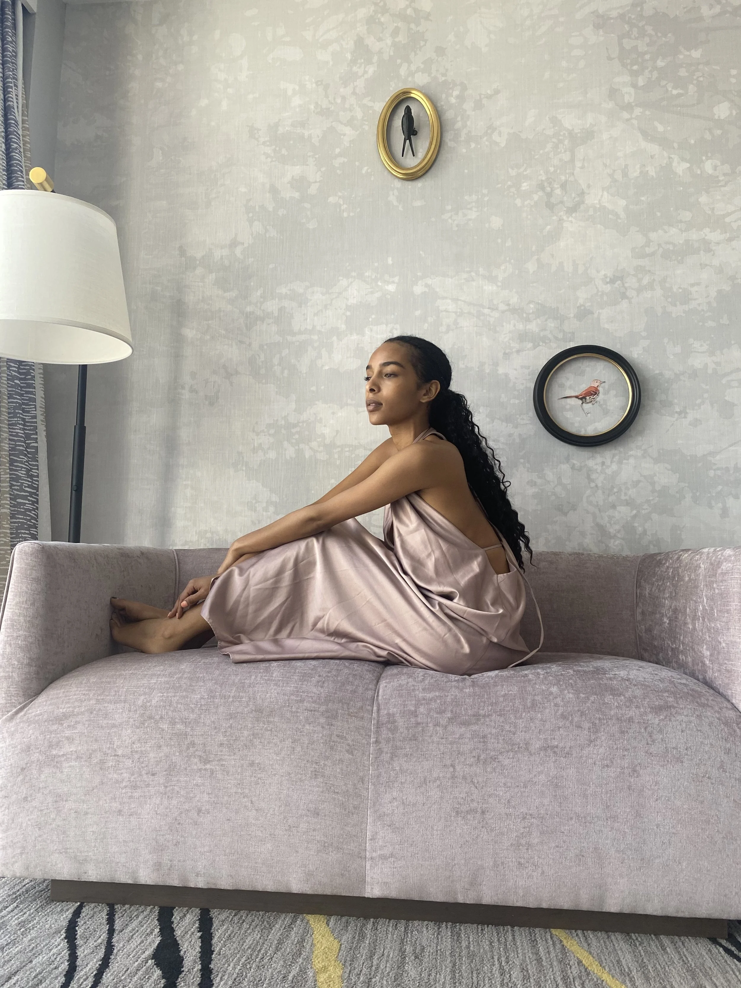 A woman sitting on a pinkish sofa with a light-colored, textured wall in the background. The woman is wearing a silky, pale pink dress and is posed with her left arm resting on her knees, gazing to her right. The wall behind her has two framed bird illustrations, one with a black frame and the other with a gold frame. To the left, there is a white lamp with a shade and a window with patterned curtains. The floor has a rug with a striped design.