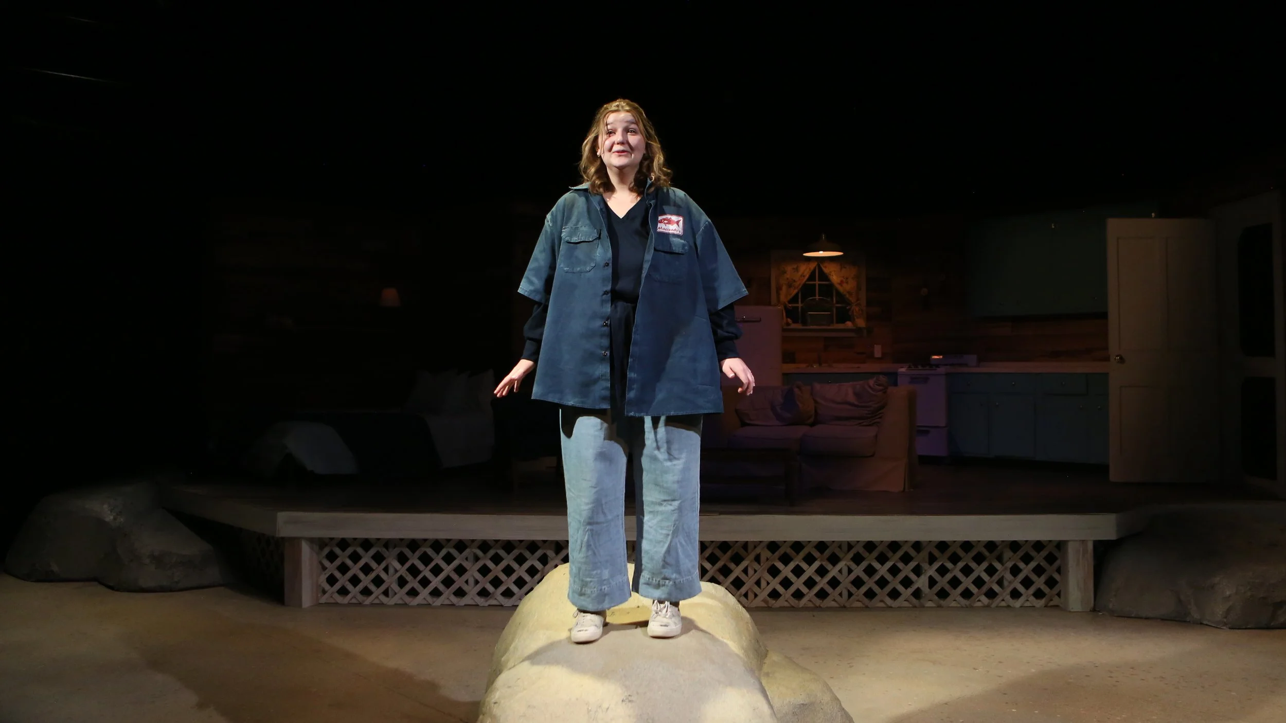 Young woman standing on a large rock on stage in a cozy, rustic living room setting, wearing a dark jacket, black shirt, and light jeans.