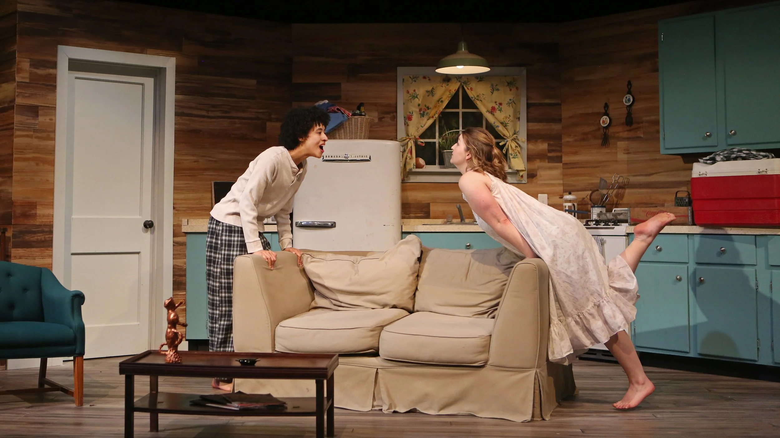 Two women are engaged in a lively conversation in a cozy kitchen and living room stage production setting, with one woman leaning on a sofa and the other standing near it, both displaying animated expressions.