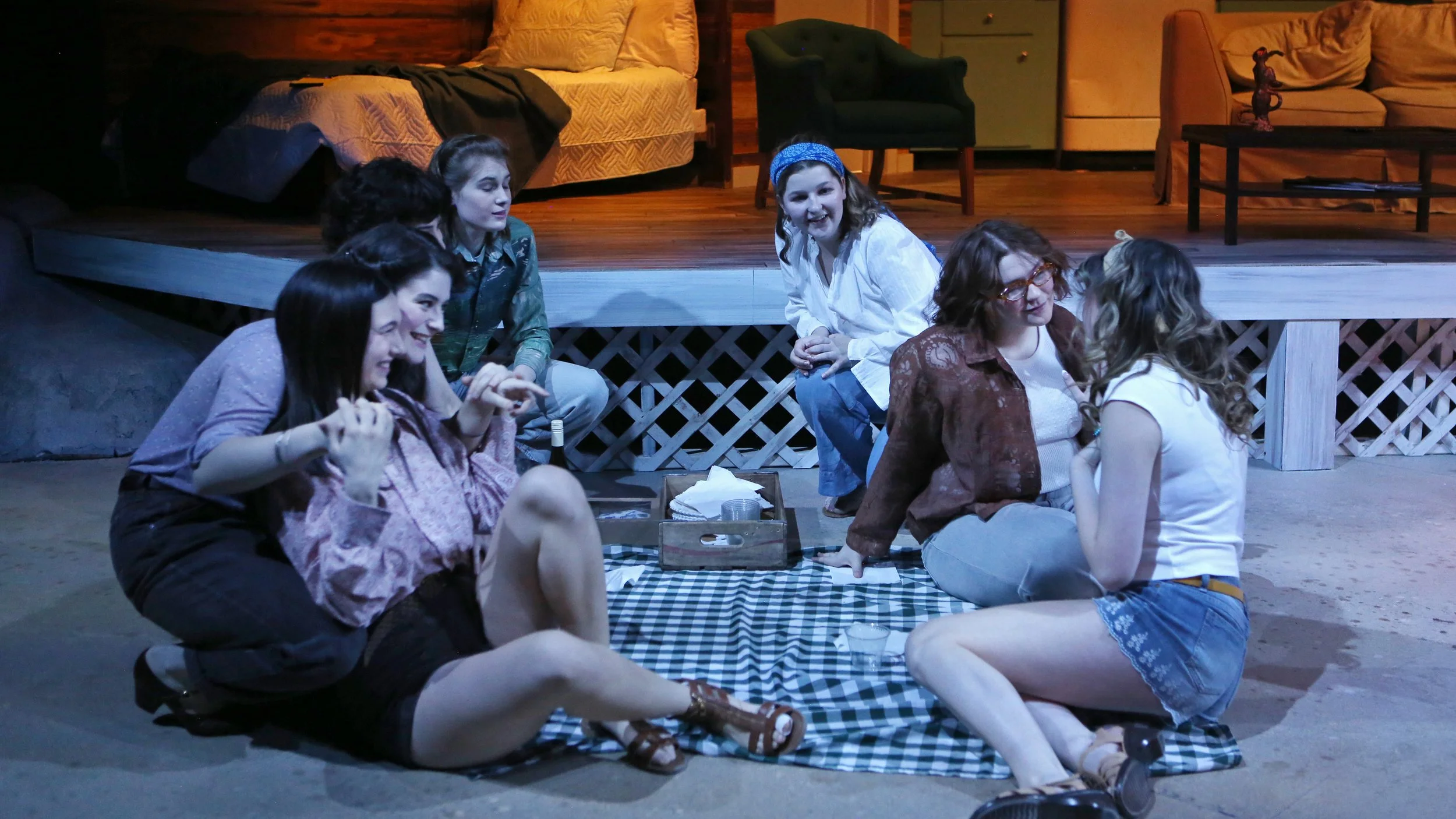 A group of seven women sitting on a checkered blanket and on the floor in front of a stage, engaging in conversation and sharing a moment of laughter. The stage behind them is set with furniture including a bed, armchair, and sofa, suggesting a cozy,