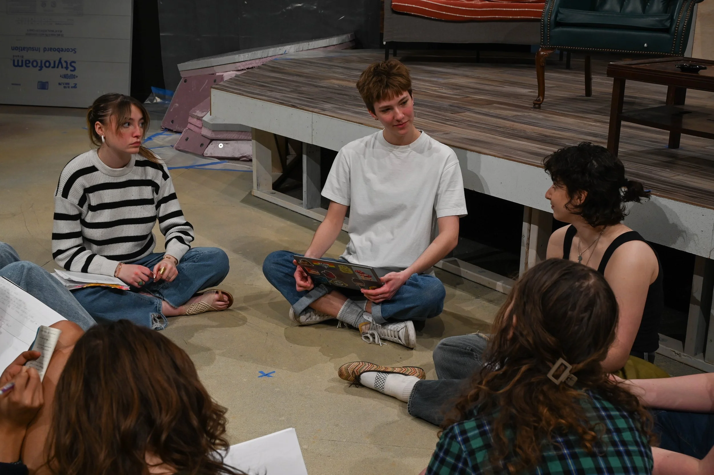 Group of actors sitting on the floor in a circle engaged in conversation, with a stage in the background.