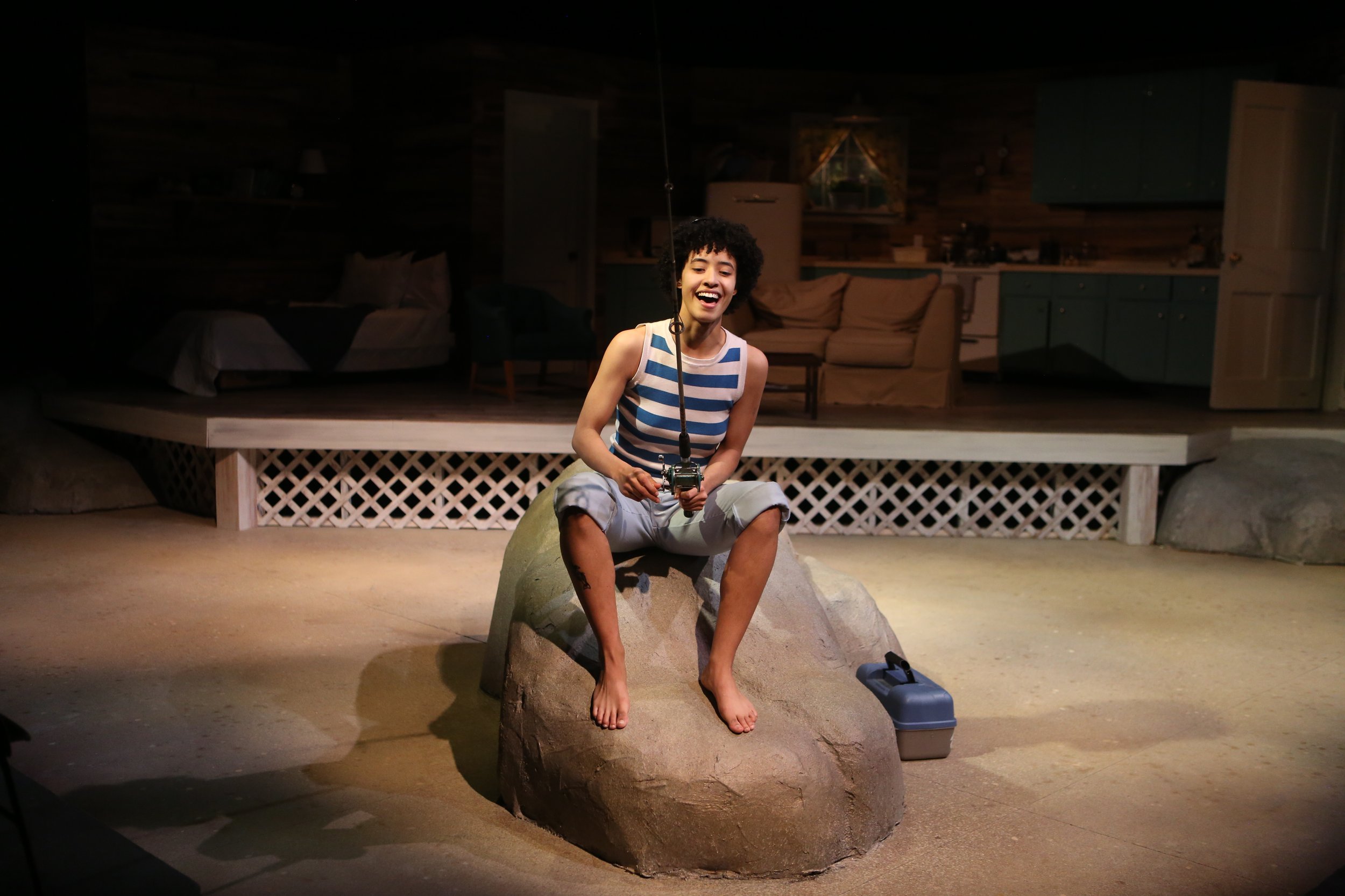 A young person with curly hair wearing a striped tank top and shorts, sitting on a large rock inside a cabin, smiling and holding a fishing reel. The background features wooden paneling, a couch, a bookshelf, and kitchen cabinets.