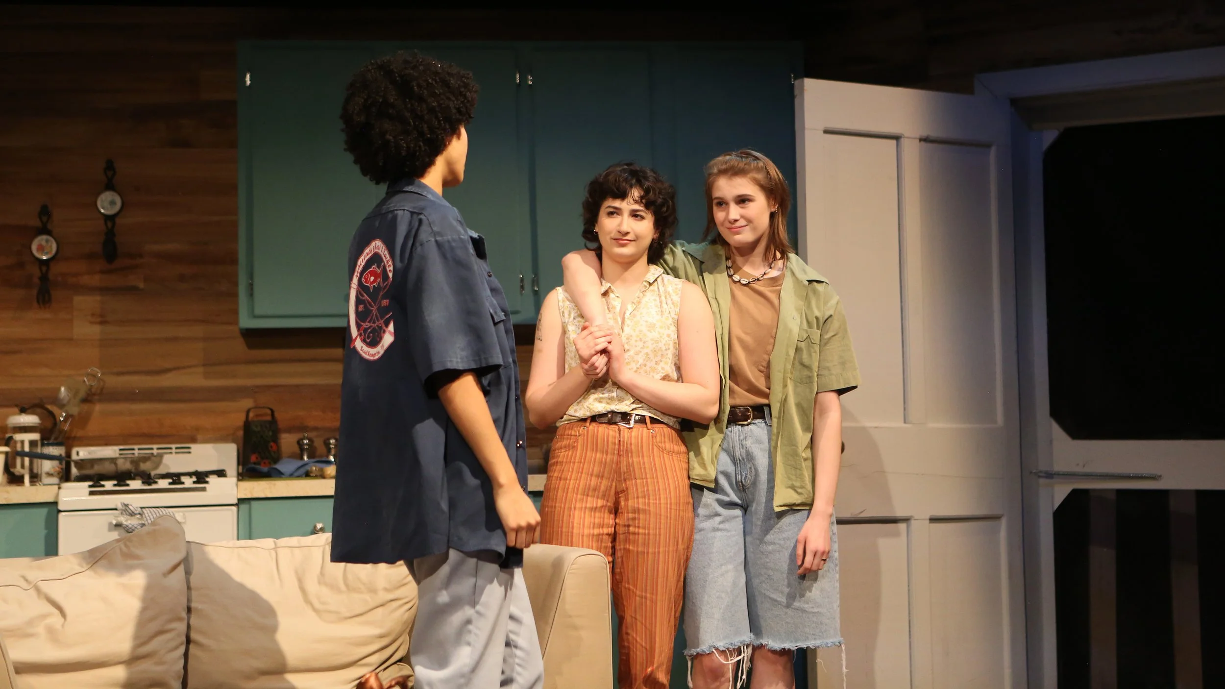 Three young women and one young man are standing in a kitchen, engaging in conversation. The man has curly hair and is wearing a dark shirt with a patch on the sleeve. Two women are facing him, one with curly hair, wearing a sleeveless floral top and