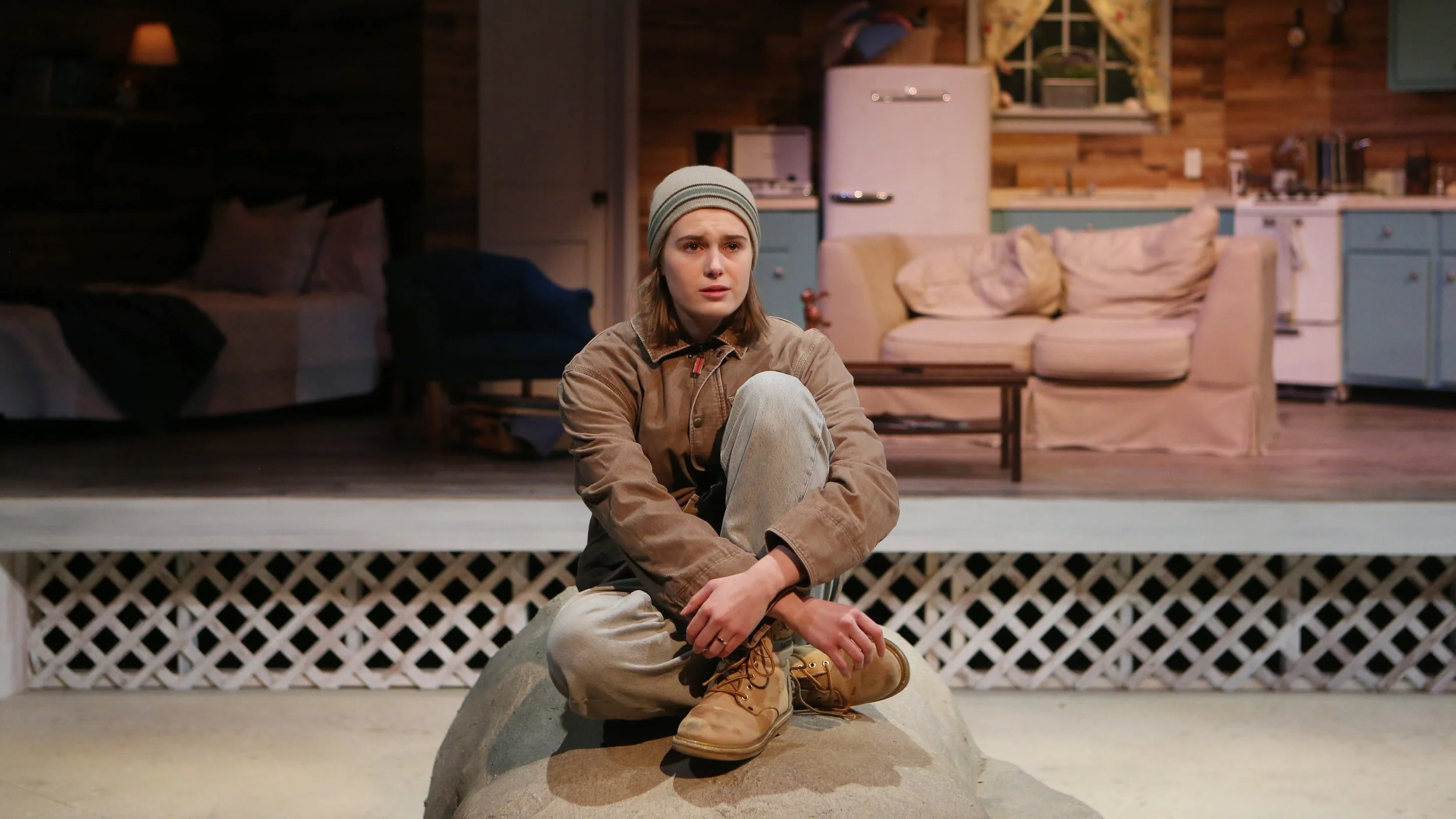 A young woman in a beanie and casual clothing sitting on a rock in front of a house with a cozy interior visible through a window.