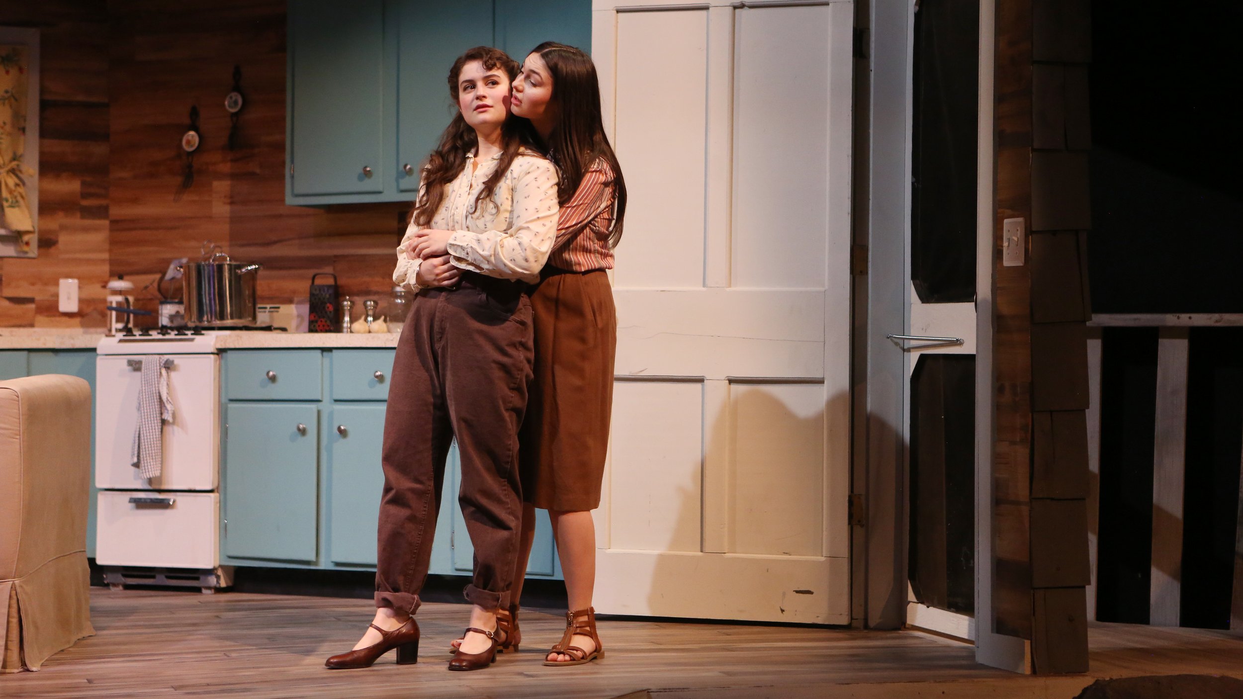 Two women standing close together in a kitchen, one whispering into the other's ear. The woman on the left has curly brown hair, wearing a light-colored blouse and brown pants. The woman on the right has straight dark hair, wearing a striped shirt an