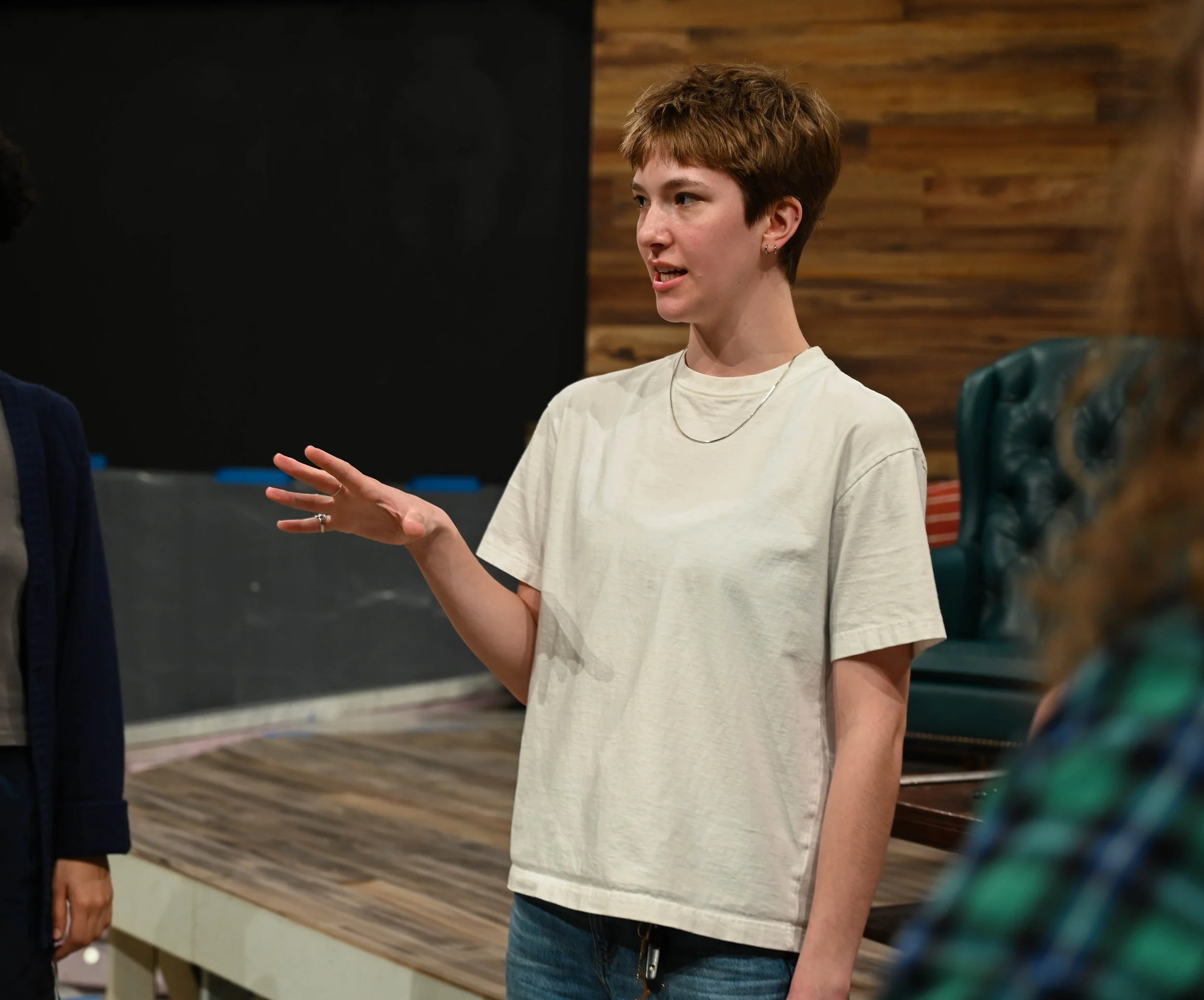 A young woman with short hair and earrings gestures with her hand while speaking in front of the set for a theater production.