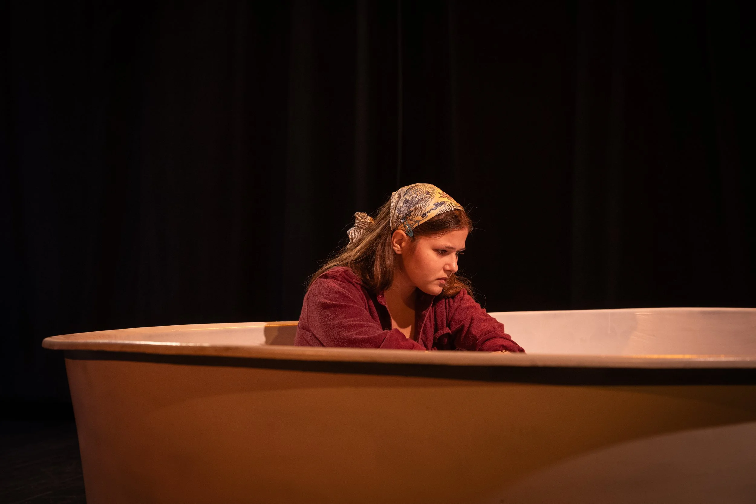 A young woman with long brown hair wearing a patterned headscarf and red jacket sitting inside a large, round, wooden tub or basin on stage against a black background.