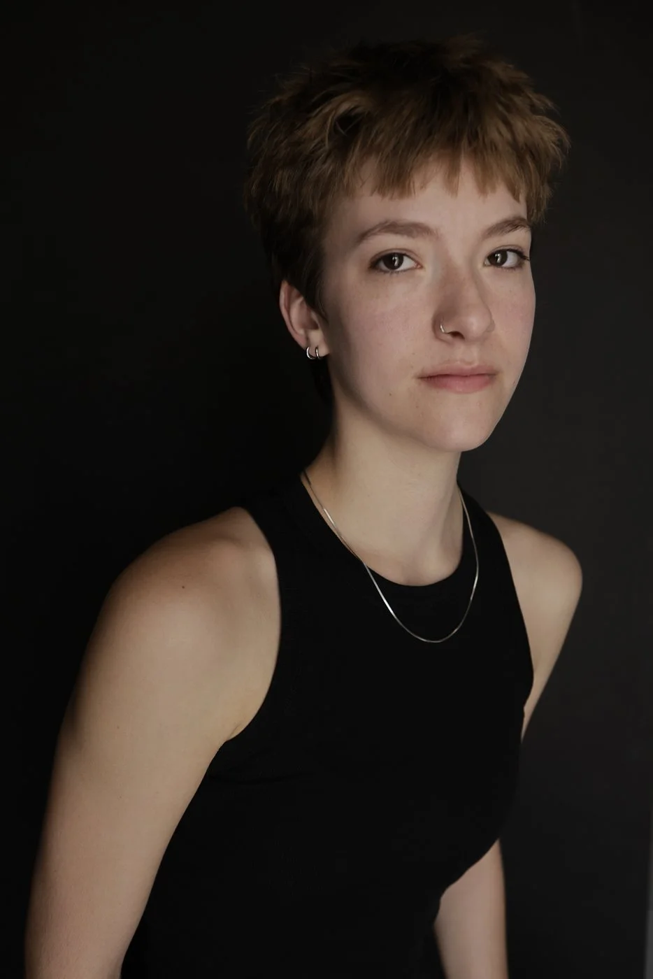 Portrait of a young woman with short, reddish-brown hair, wearing a black sleeveless top, silver necklace, and earrings, against a dark background.
