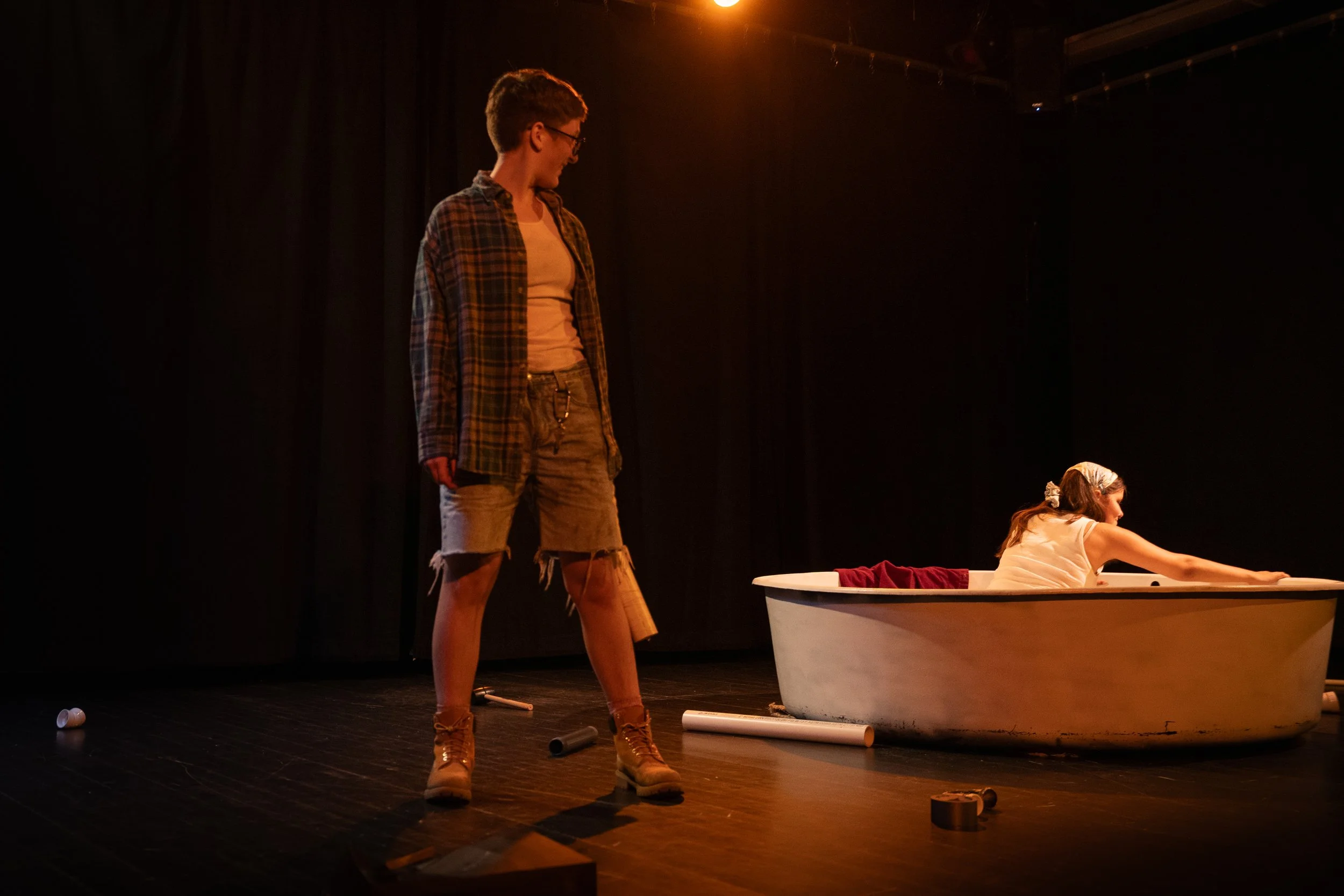 A theater stage with two young actors performing a scene. One actor, a boy in a plaid shirt, ripped shorts, and boots, stands near a bathtub. The other actor, a girl in a white dress with a bow in her hair, is reaching into the bathtub, which has a r