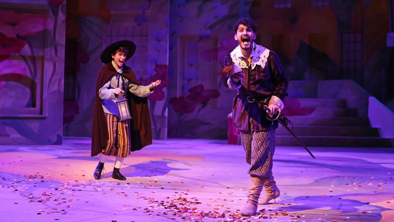 Two actors performing on stage with colorful, theatrical scenery; one actor is holding a lantern, and the other is holding a sword, both smiling and engaging with the audience.