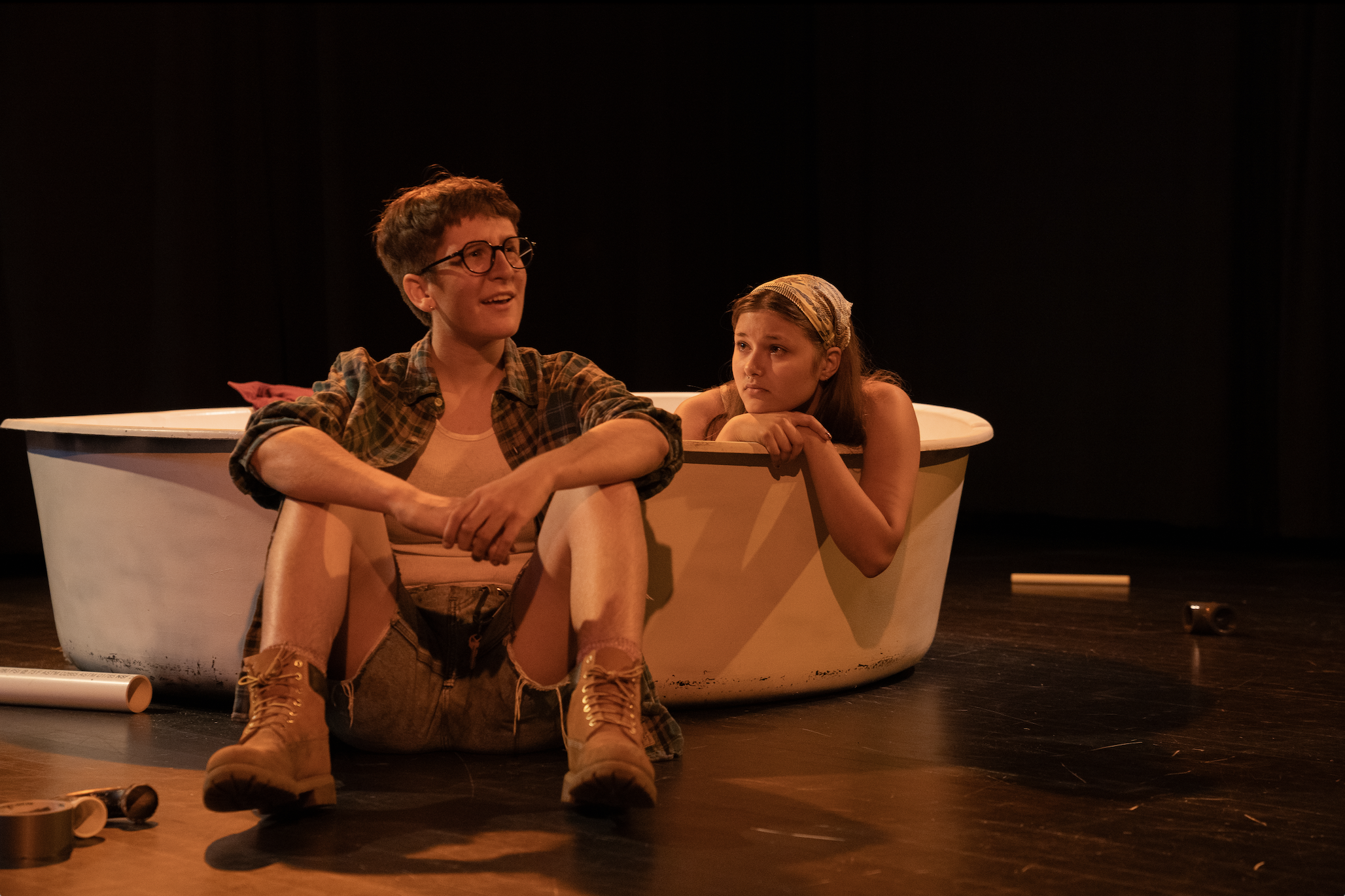 Two young actors sitting in a white bathtub on a stage, with theatrical props on the floor, during a performance.