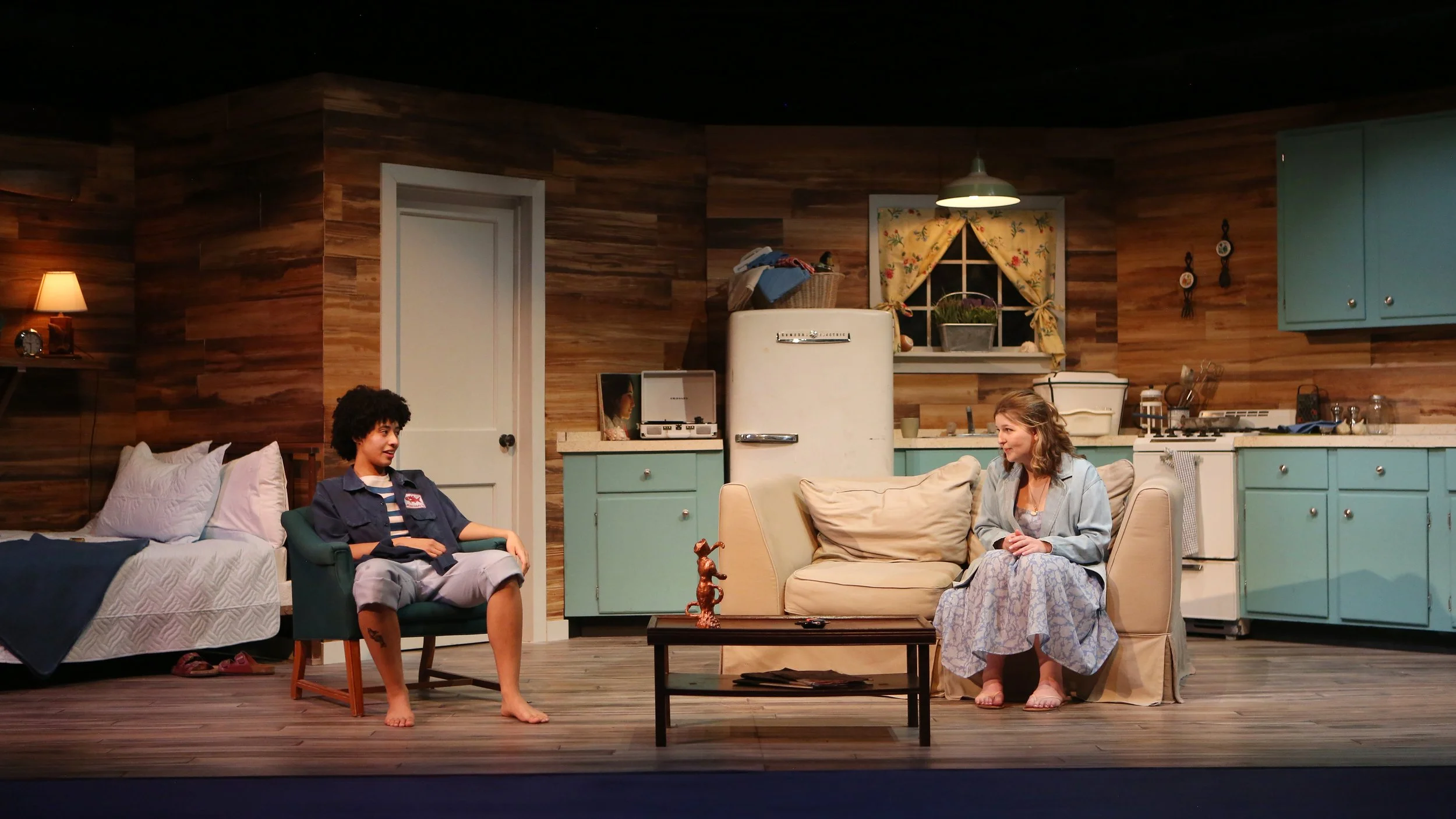 Two women are sitting in a cozy vintage-style kitchen with wooden walls, thee women are talking relaxed. One woman is sitting on a beige sofa, the other on a green armchair, and a small wooden coffee table is between them. The kitchen features pastel