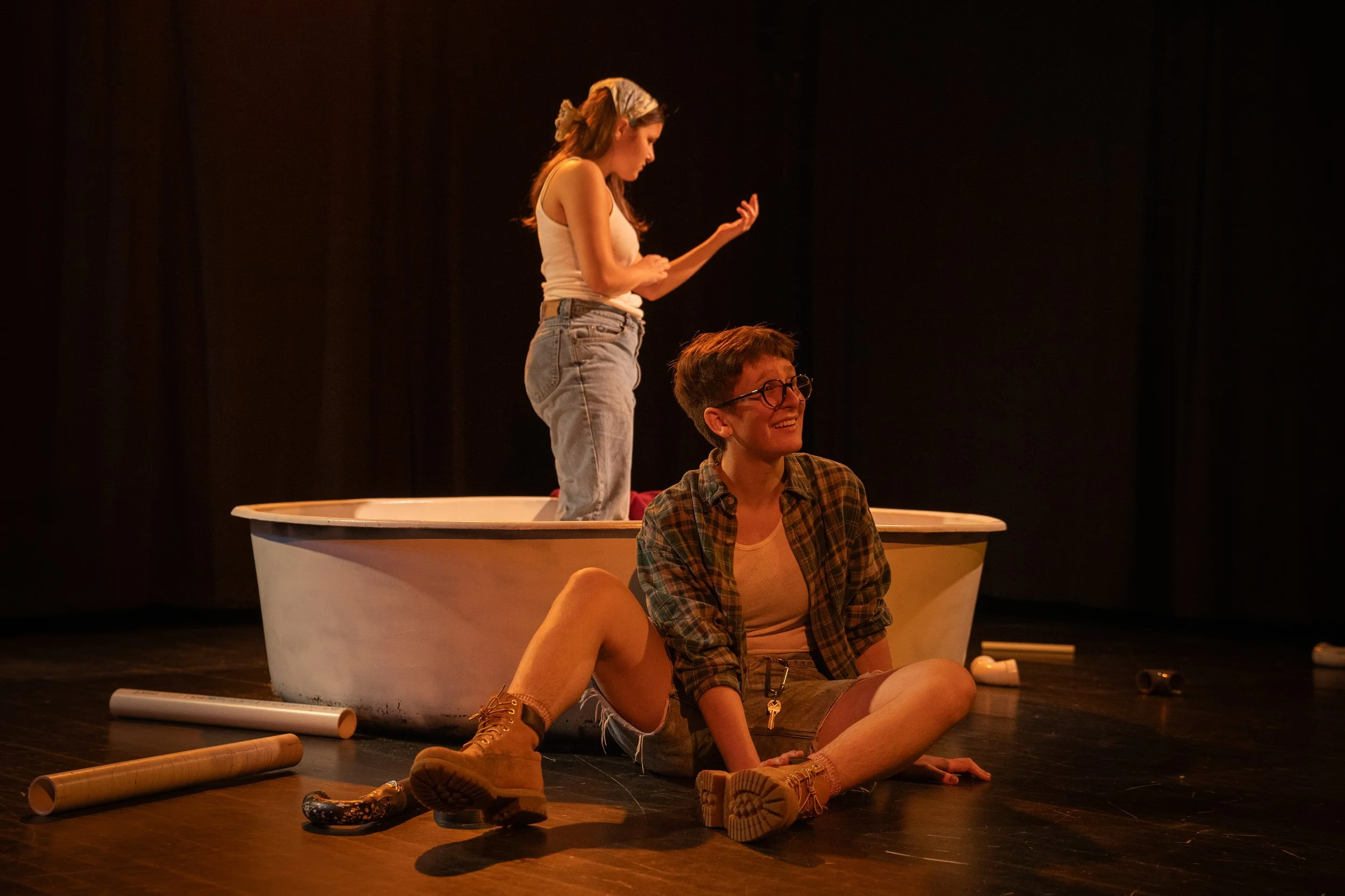 A young woman standing on a bathtub with her right hand raised, and another young woman sitting on the floor in front of her, smiling. The scene appears to be a theatrical performance with props scattered on the floor.