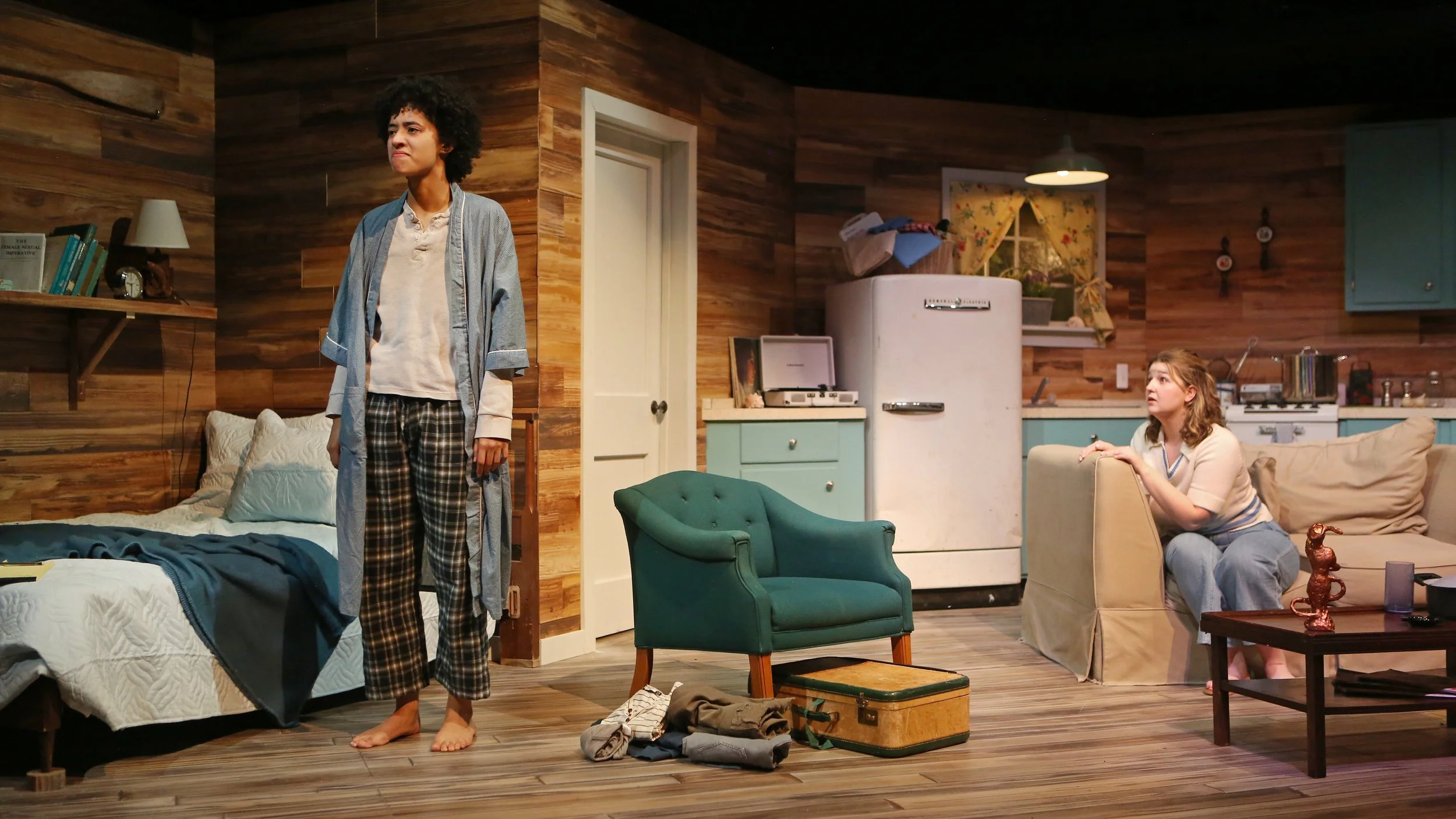 A scene from a stage play set in a cozy, vintage-style kitchen and bedroom. A man with curly hair, wearing pajamas, stands barefoot in front of a bed with a surprised or upset expression. A woman with curly hair, dressed casually, sits on a beige arm