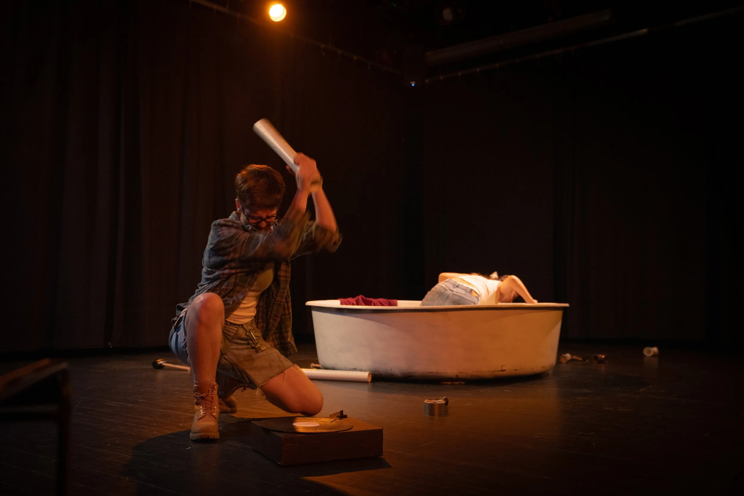 A person with short hair, glasses, and wearing a plaid shirt is kneeling on a stage, holding a long object above their head. There is a white bathtub behind them, with someone lying face down inside it. The scene appears to be a theatrical performanc
