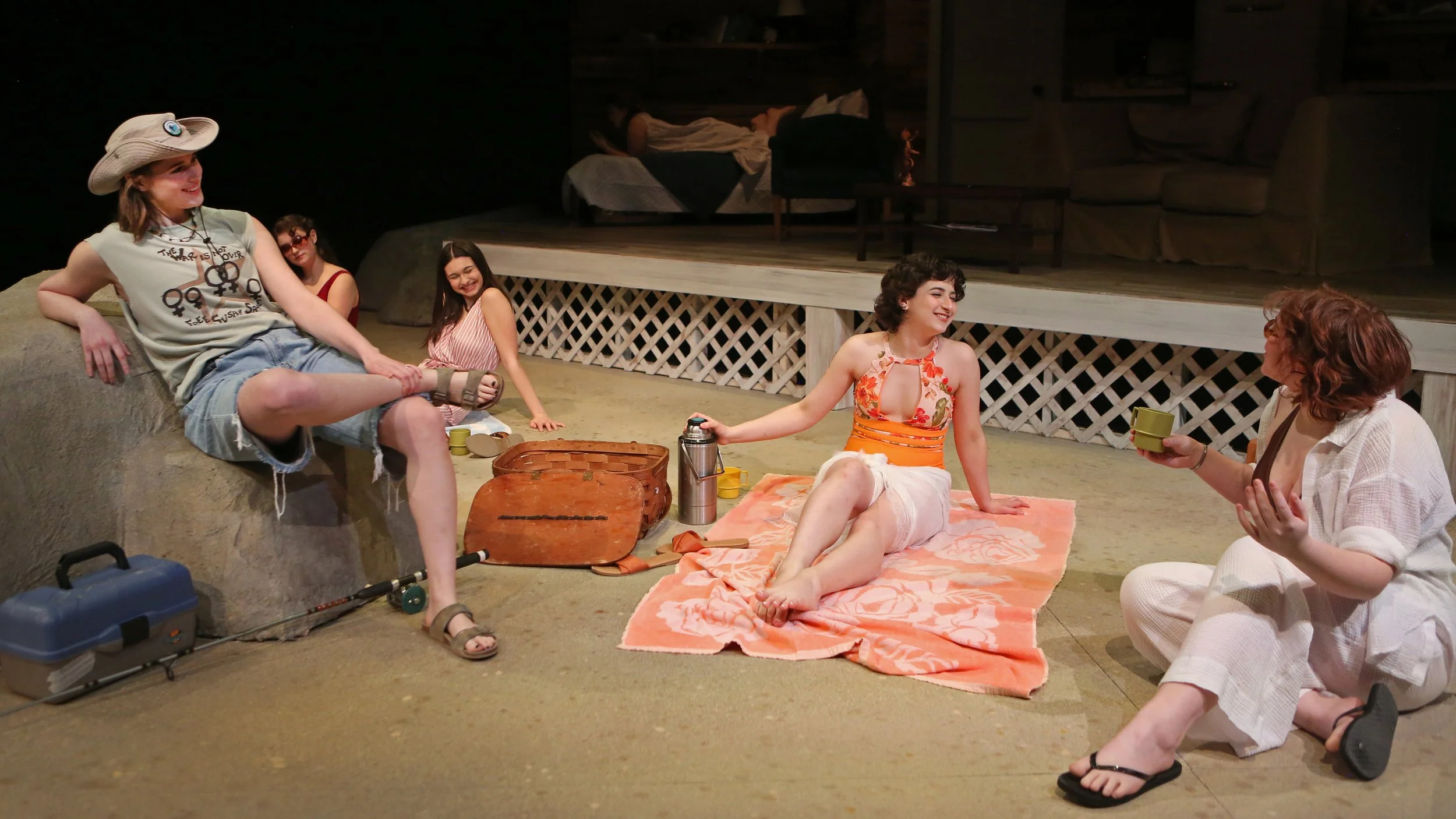 A group of six young women sitting and relaxing on the floor of a stage set designed to look like a cozy living room. They are smiling, chatting, and enjoying beverages. Some are seated on blankets or cushioned surfaces, with props such as a picnic b