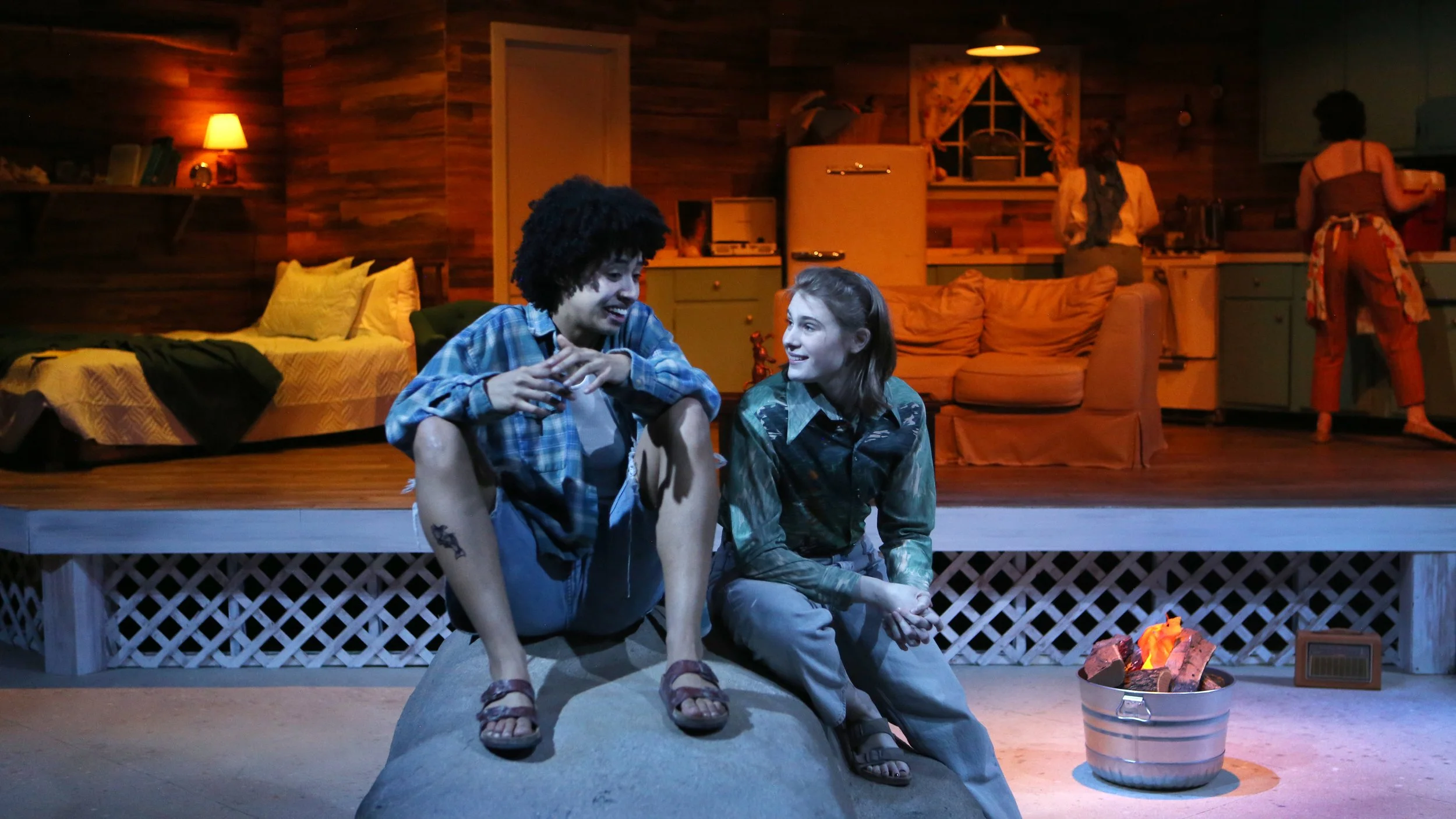 Two young people sitting on a raised porch in front of a cozy, rustic wooden cabin interior. The young man has curly hair and is wearing a blue plaid shirt, while the young woman has straight hair and is wearing a camouflage jacket. They are smiling 